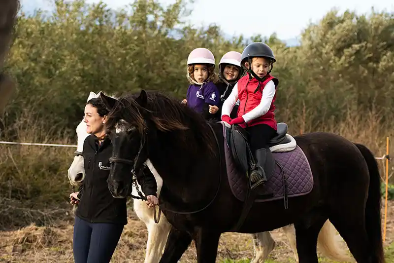 Three young children wearing helmets ride a dark pony guided by a woman outdoors.