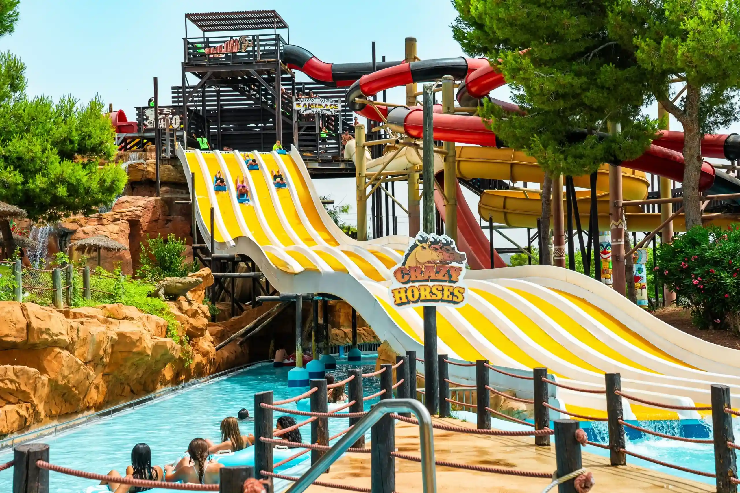 People racing down a large yellow multi-lane water slide at a theme park with other slides behind.