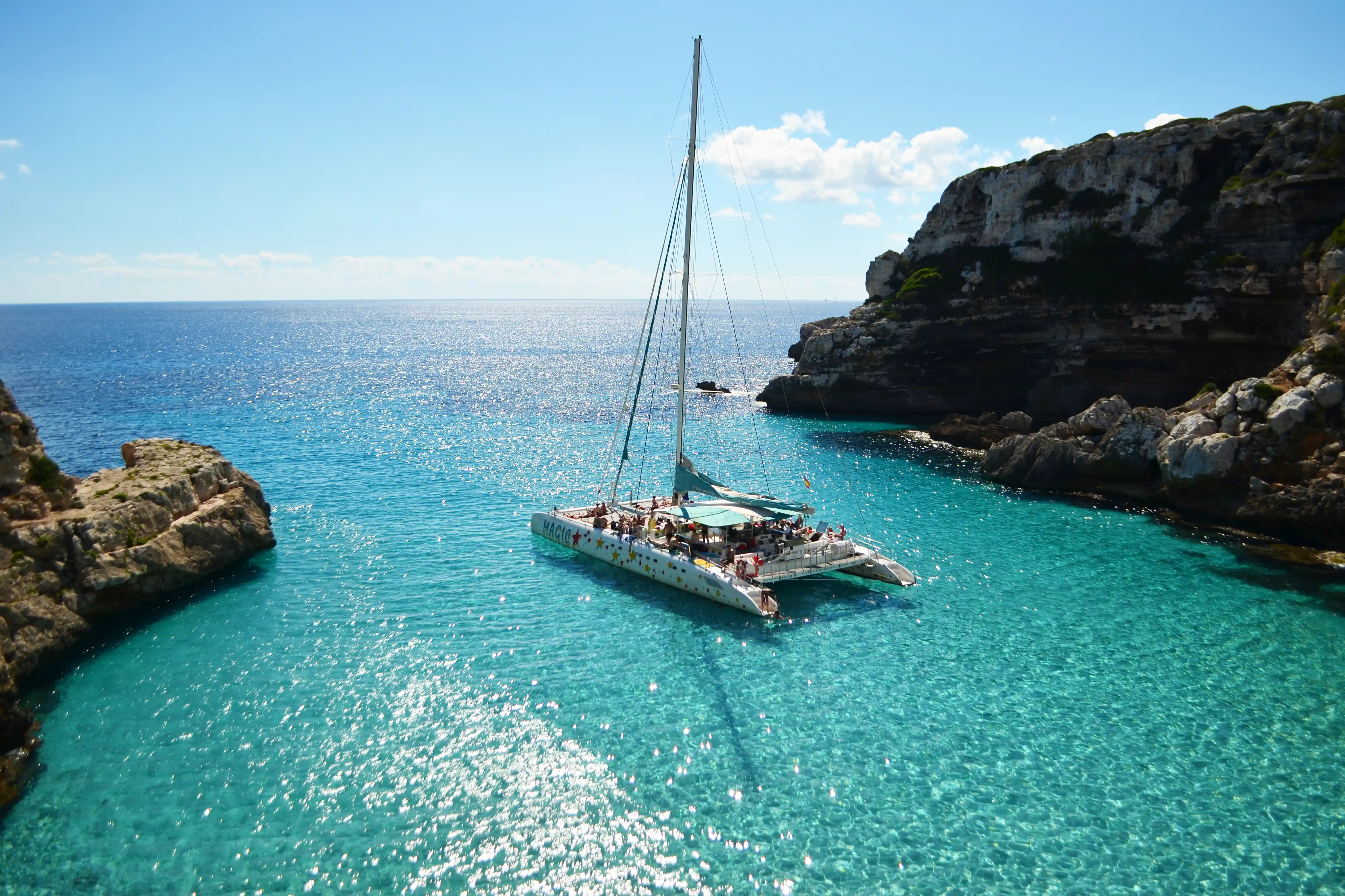 A large catamaran with tourists anchors in a beautiful turquoise bay surrounded by rocky cliffs on a sunny day.