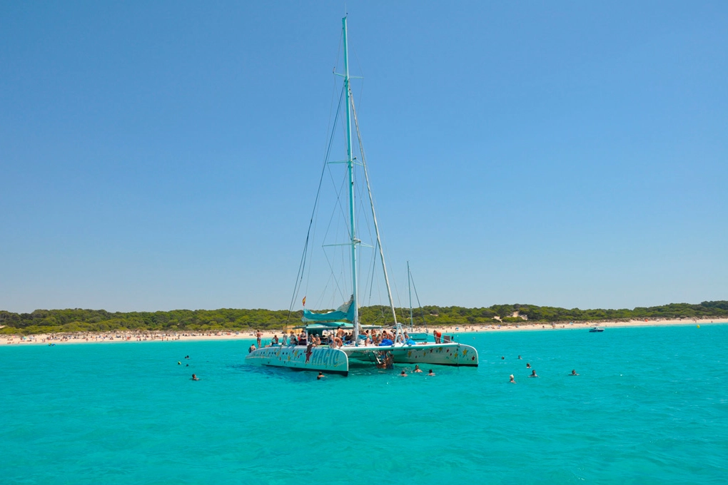A large catamaran sailboat anchored in the turquoise sea with tourists swimming and sunbathing near a white sand beach in Mallorca.