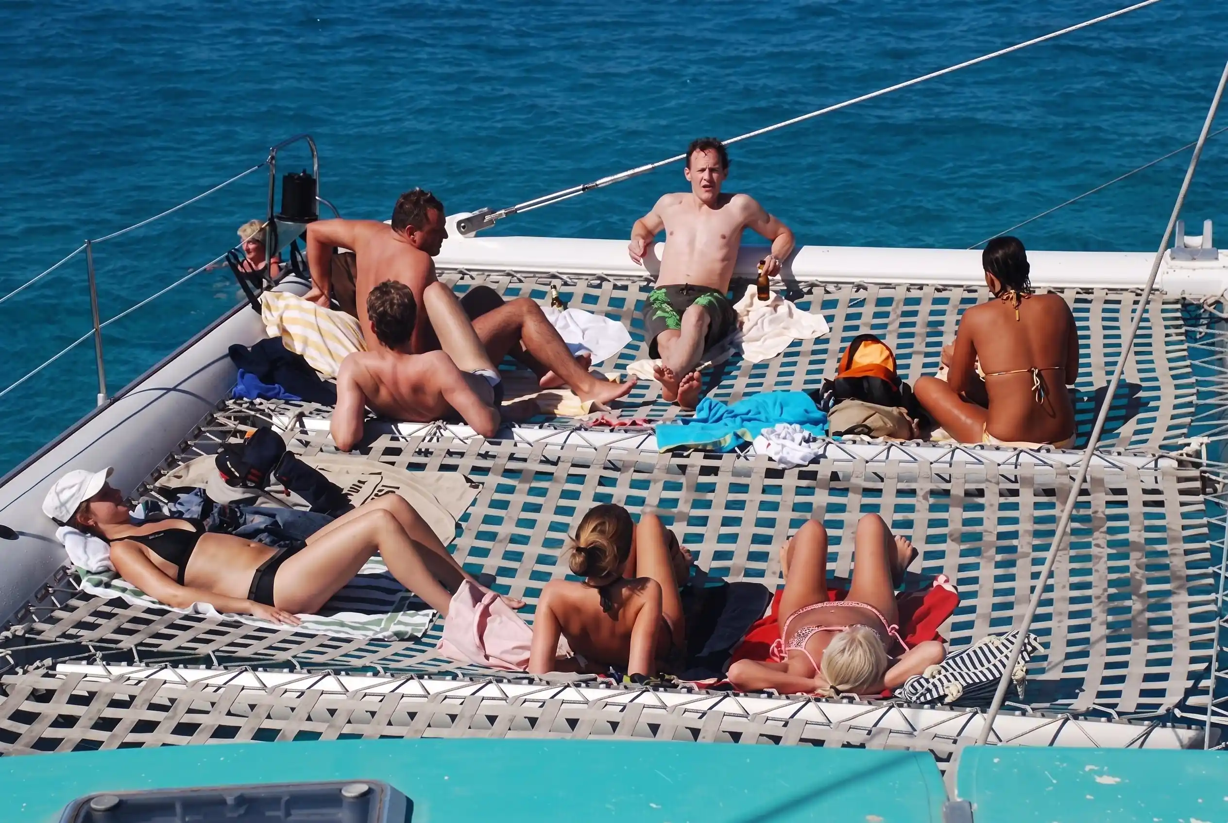 A group of people relaxes and sunbathes on the front net of a large catamaran sailing on the deep blue sea.