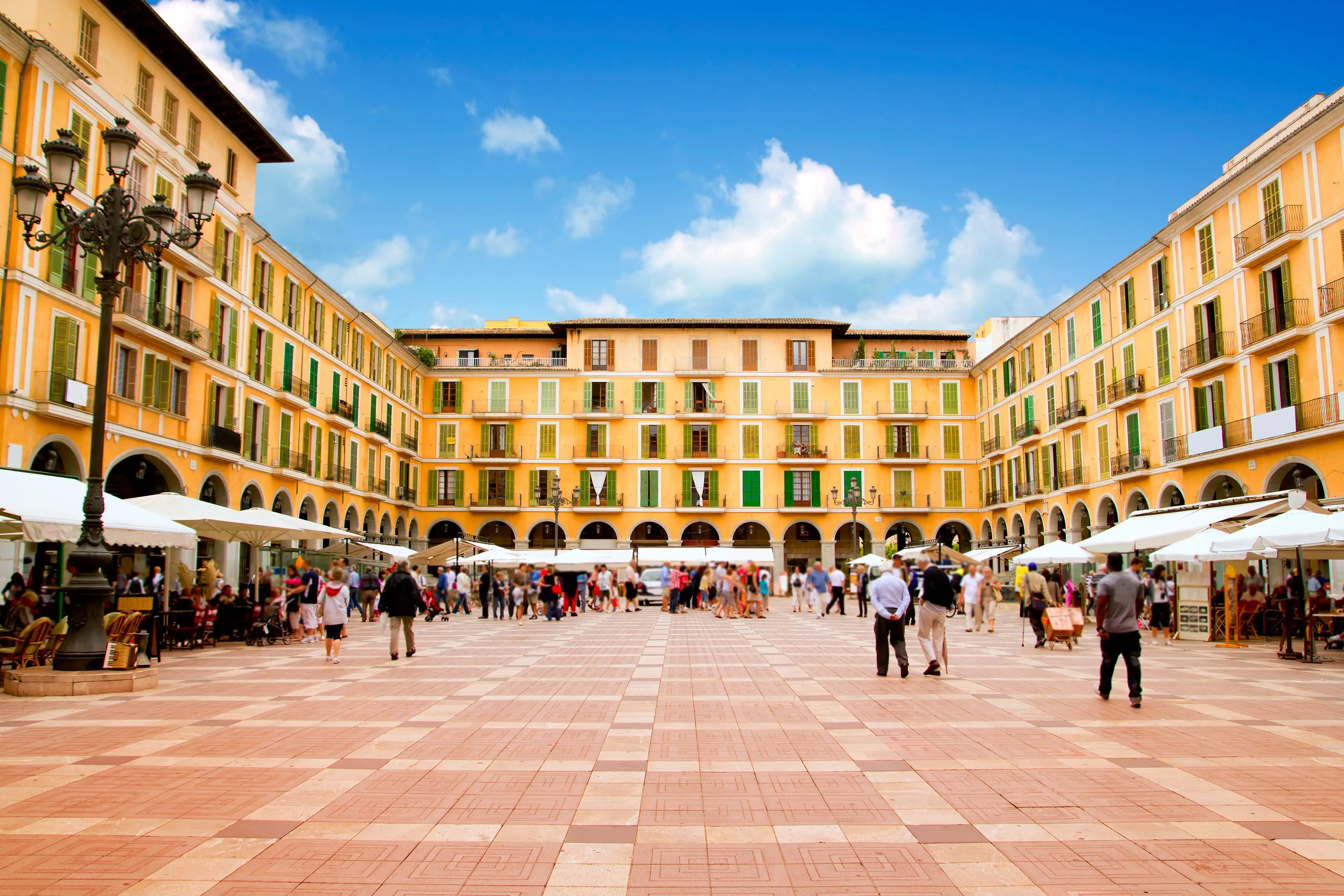 A bustling public square surrounded by traditional yellow buildings with green shutters under a partly cloudy sky.