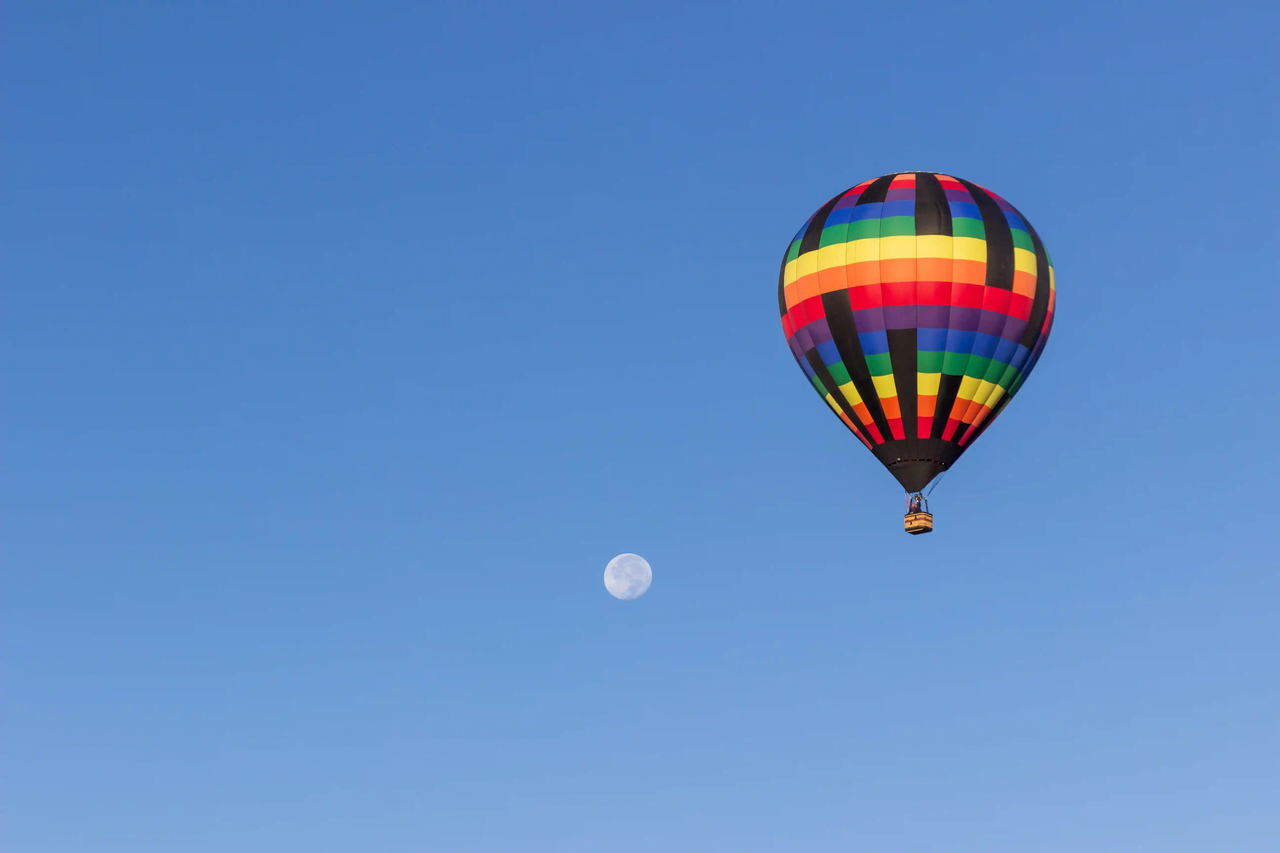 A rainbow-colored hot air balloon flying in the blue sky next to a visible daytime moon.