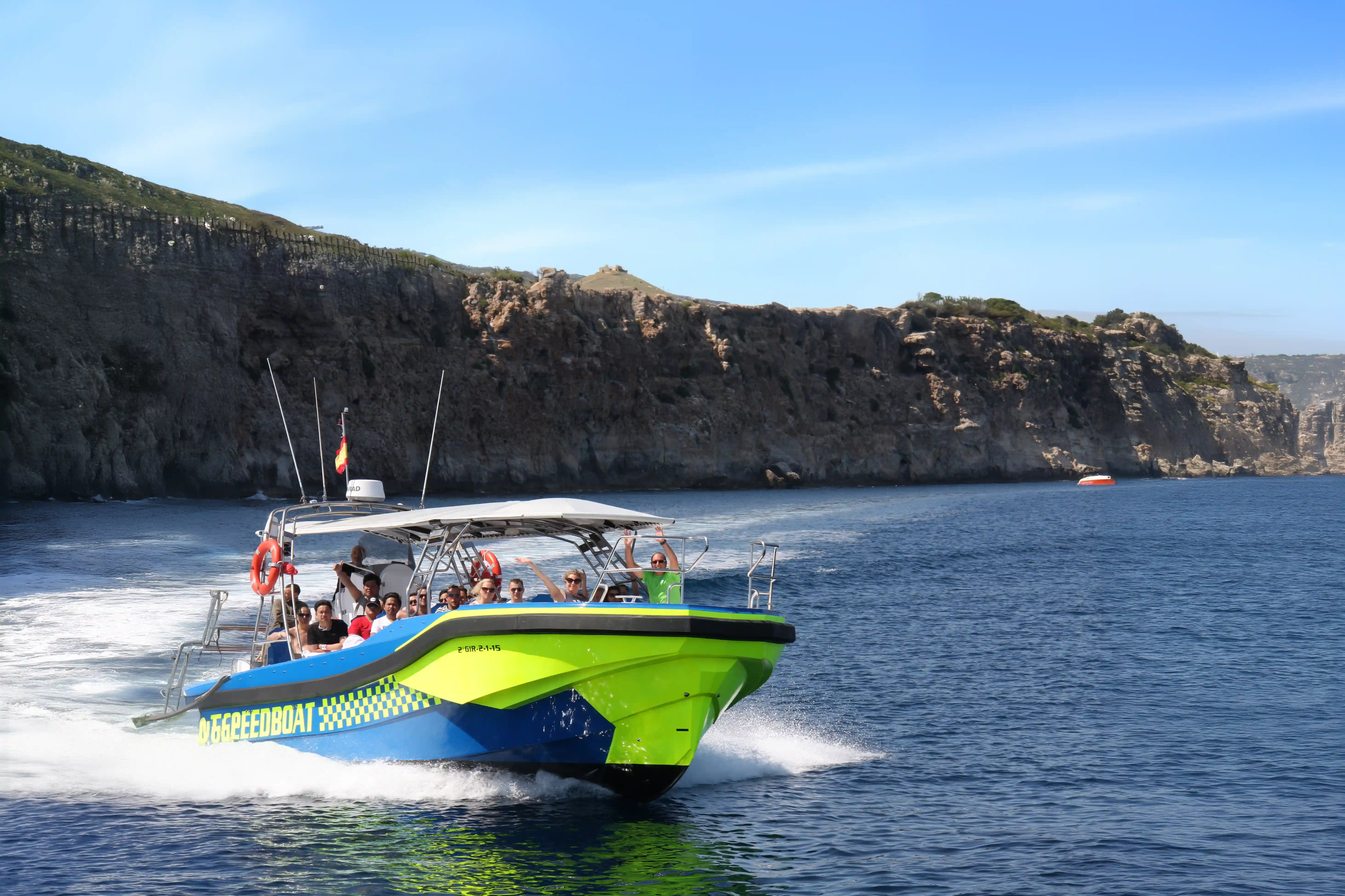 A bright green and blue speedboat carrying passengers close to high, rocky coastal cliffs.