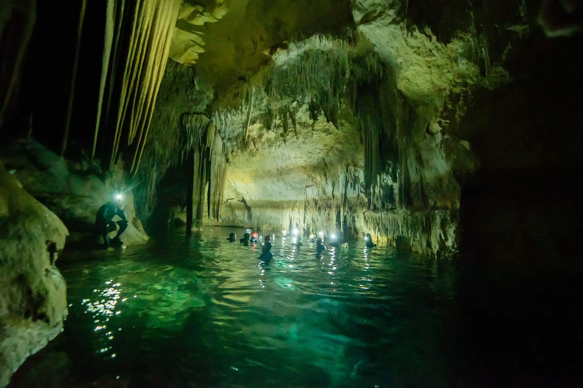 People swimming with flashlights inside a dark cave with a subterranean turquoise lake and stalactites. People swimming with flashlights inside a dark cave with a subterranean turquoise lake and stalactites.