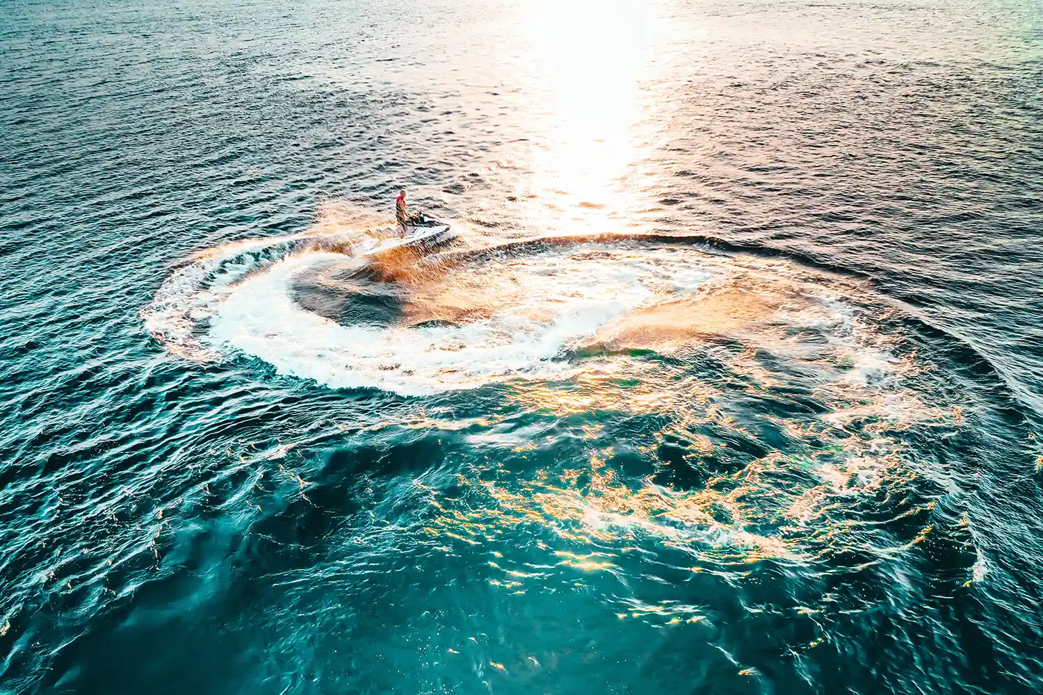 An aerial view of a jet ski turning sharply on the ocean water, creating a circular white wake at sunset. An aerial view of a jet ski turning sharply on the ocean water, creating a circular white wake at sunset.