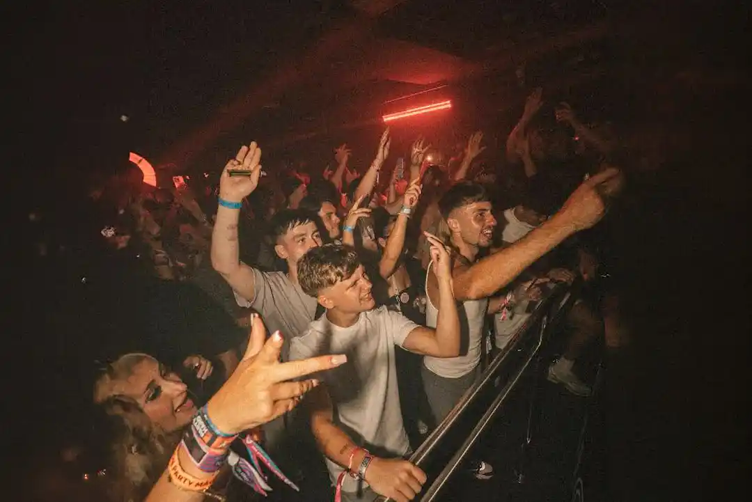 Young crowd dancing with raised hands at a concert in a dimly lit venue with red lights. Young crowd dancing with raised hands at a concert in a dimly lit venue with red lights.