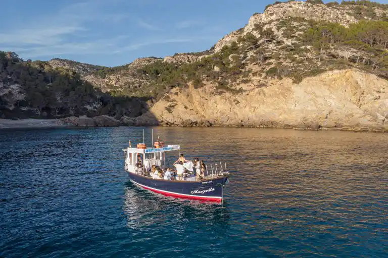 A small tour boat with passengers sails in a calm bay near high rocky cliffs during a sunny day.