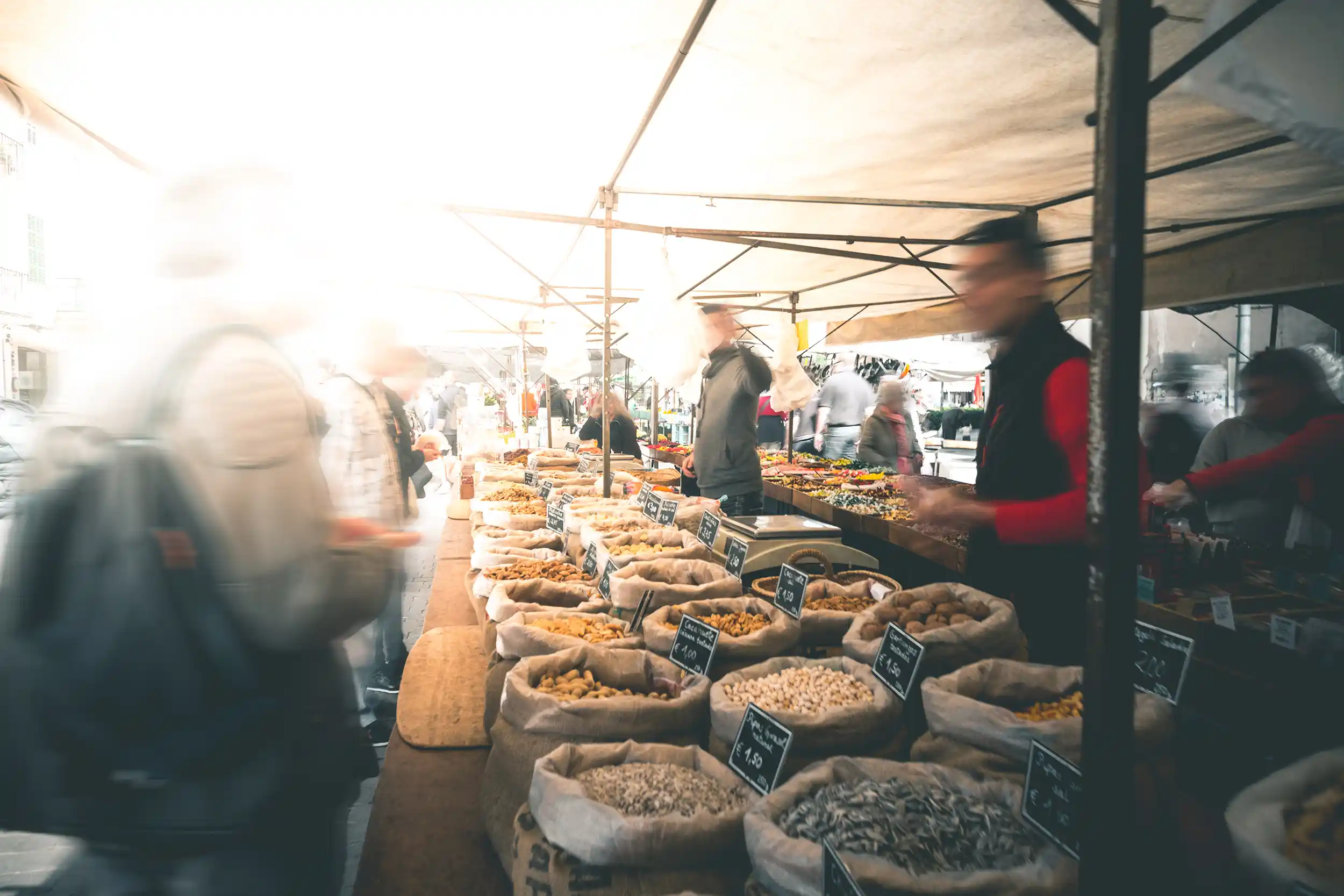 Busy outdoor local market stall grains nuts A busy outdoor local market stall with canvas sacks filled with various grains, seeds, and nuts on display as people walk by in a blurred motion.