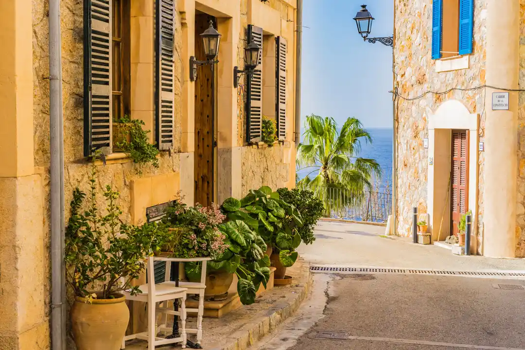 A sunny coastal street lined with historic stone buildings, green potted plants, and a view of the blue sea.