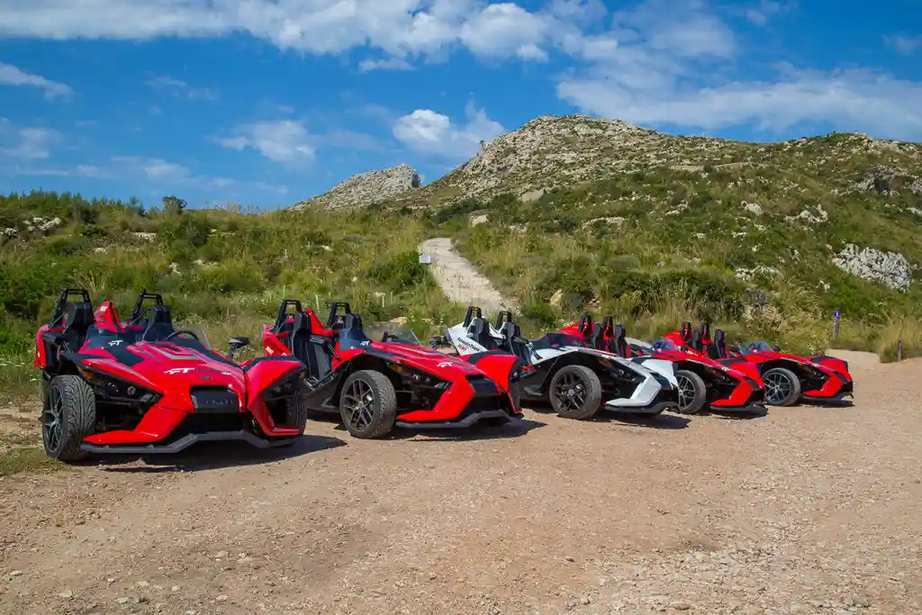 A row of red and white three-wheeled Slingshot cars parked on a dirt path with green hills behind. A row of red and white three-wheeled Slingshot cars parked on a dirt path with green hills behind.