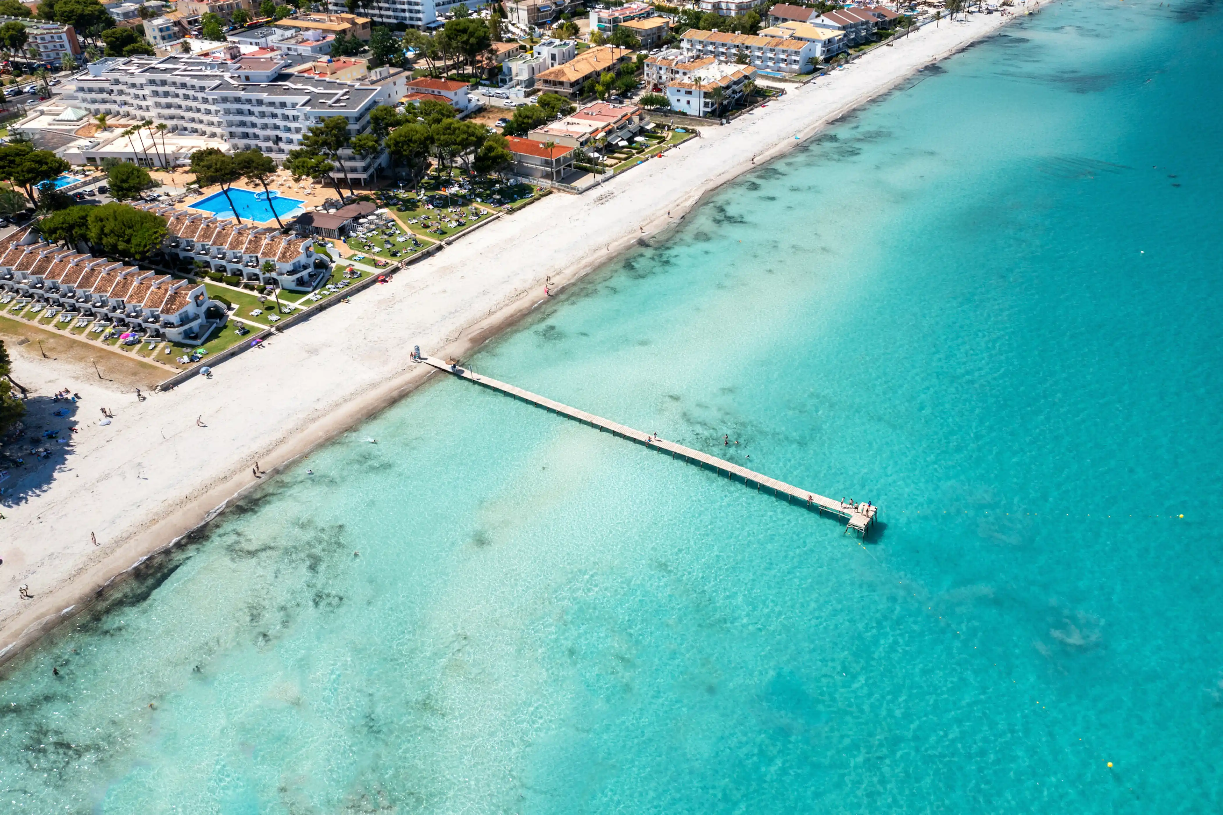 An aerial view of a long wooden pier on a sandy beach next to turquoise water and hotels in Mallorca.