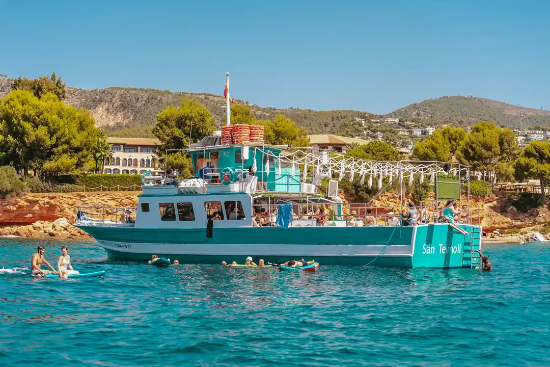 A turquoise tour boat anchors in the calm sea with people swimming nearby against a green coastal hill.