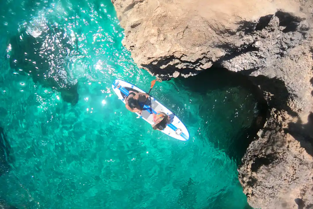 An overhead view of two people sharing a paddleboard on the clear turquoise sea next to a rocky cliff.