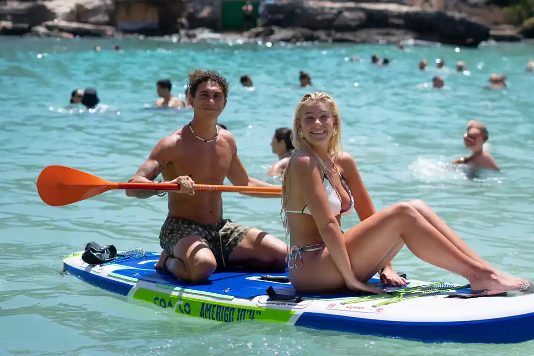 A young couple smiles while sharing a paddleboard on the clear turquoise sea during a sunny summer day.