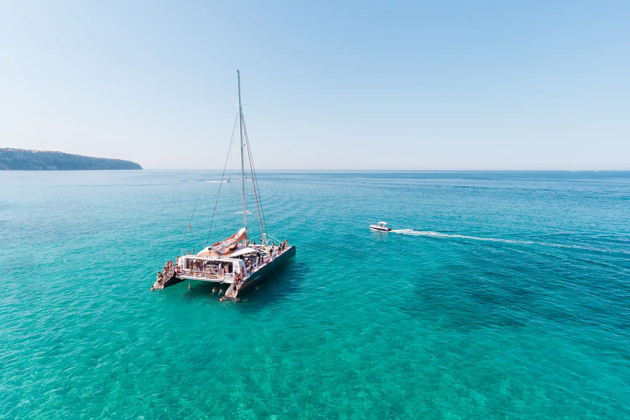 An aerial high-angle view of a large catamaran yacht filled with tourists floating on calm turquoise sea water under a clear blue sky.