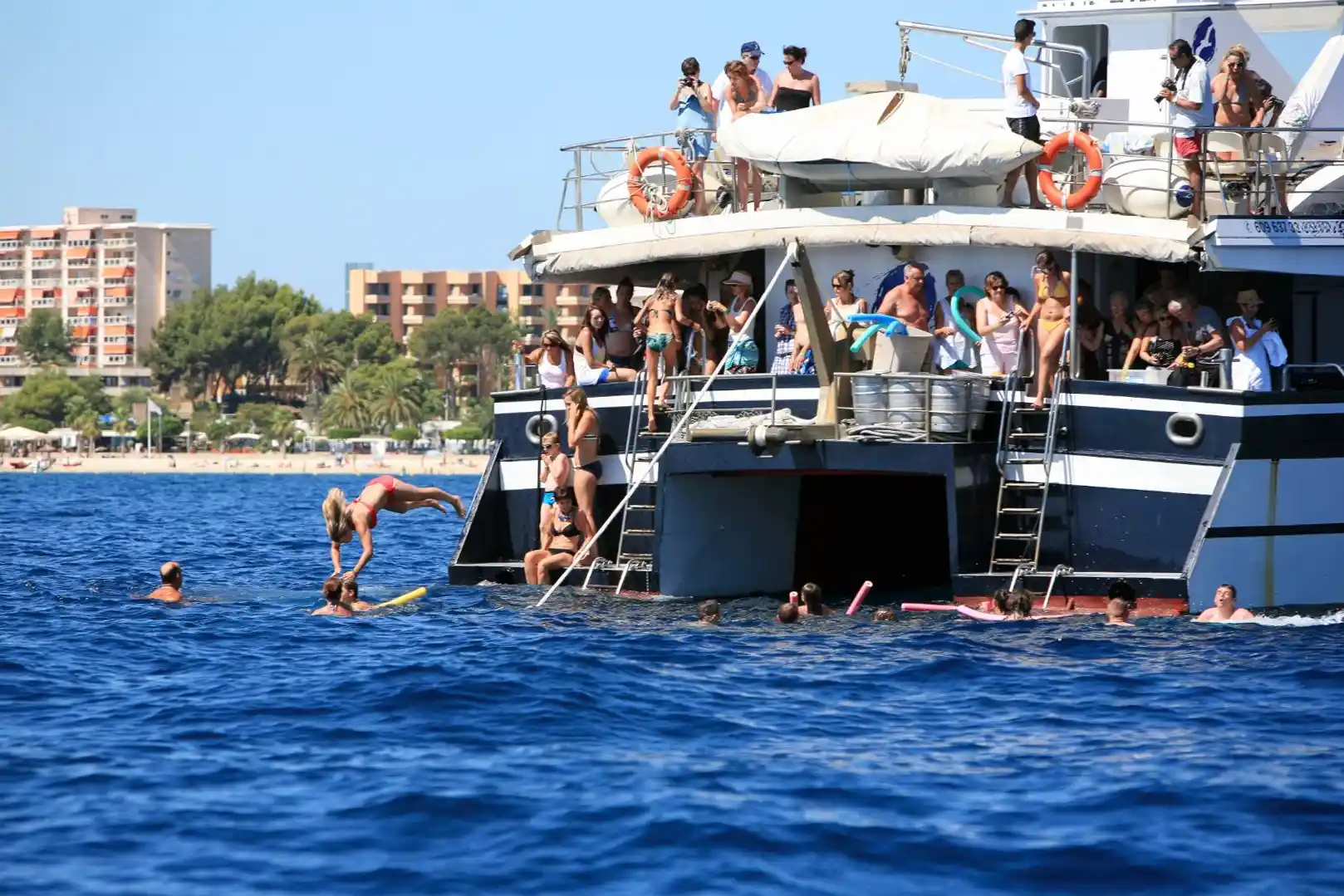 Menschen springen und schwimmen im blauen Meer vom Heck eines großen Touristen-Katamarans bei einer Sommerparty.