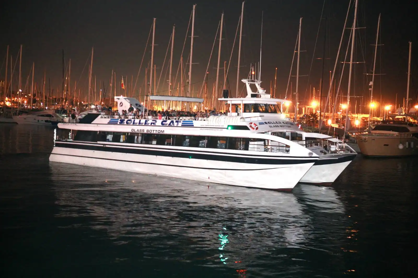 Ein großes weißes Katamaranboot liegt nachts im Hafen, umgeben von Segelmasten und Lichtern der Stadt.