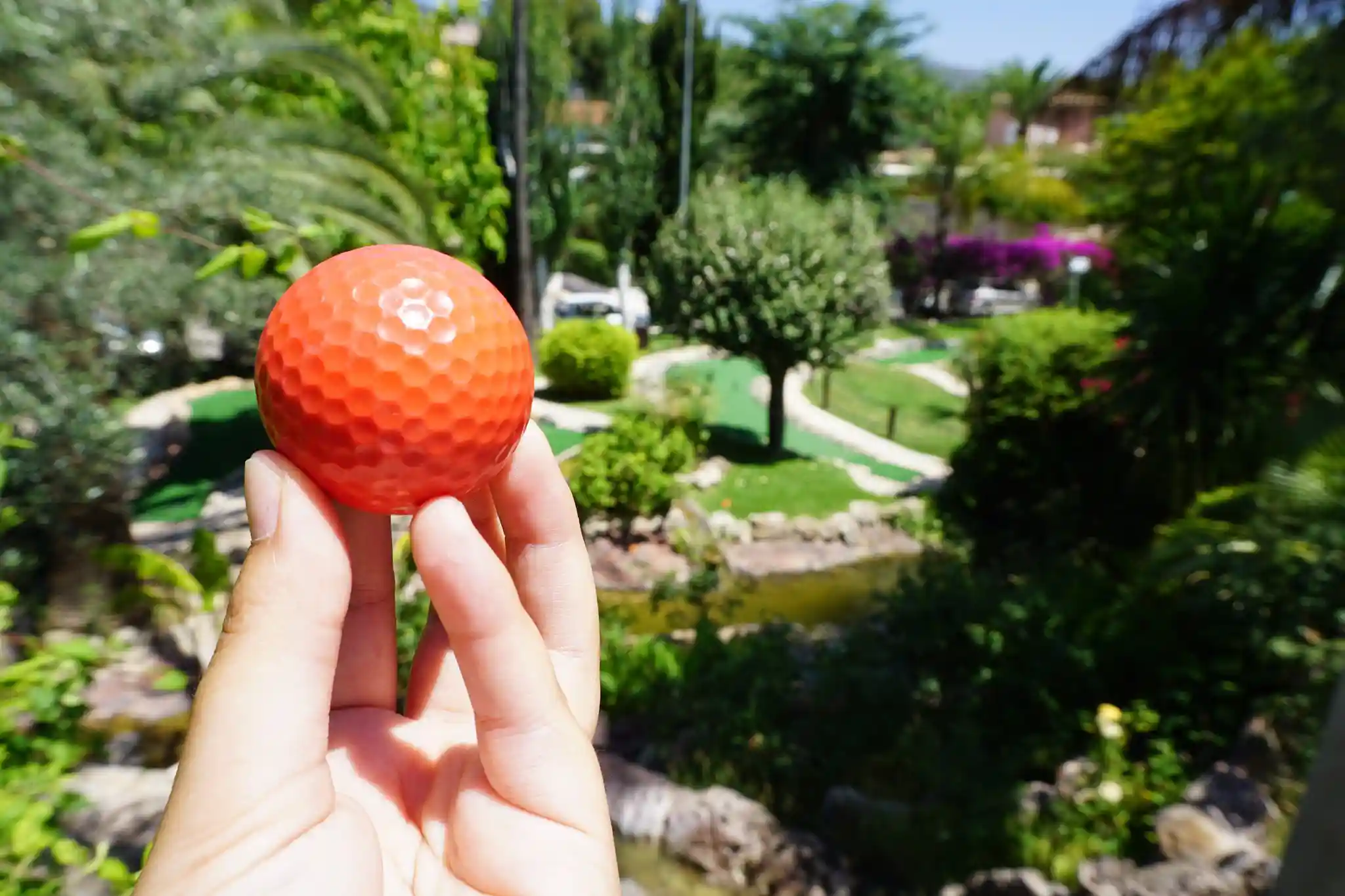 A hand holding a bright orange mini-golf ball close to the camera with a blurry green course in the background.