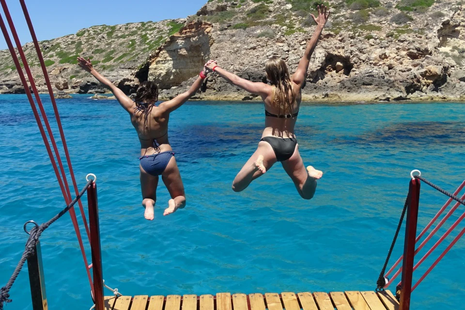 Two women in bikinis jumping hand in hand from a boat into clear turquoise sea water near a rocky coast.