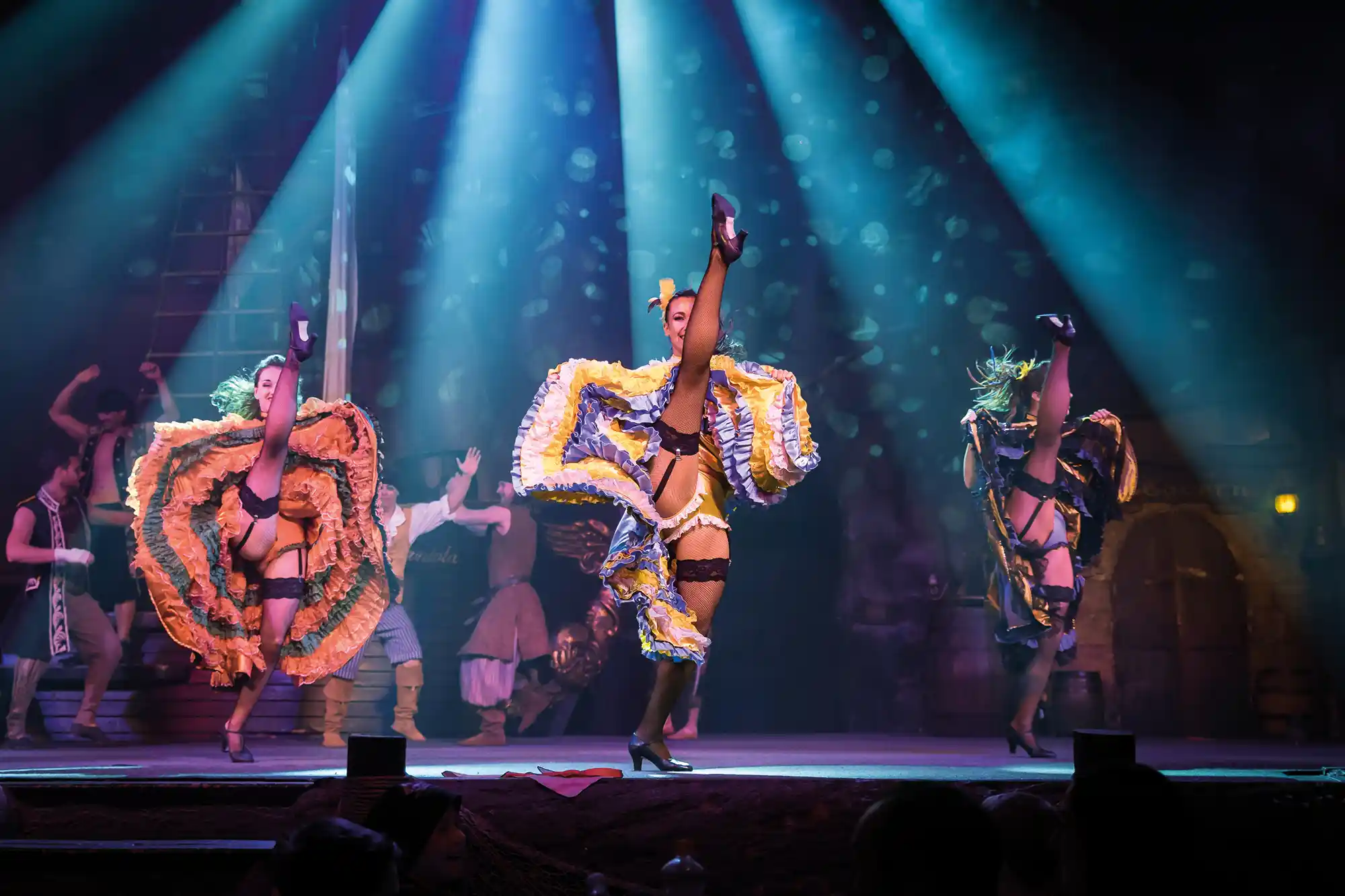 Three female dancers perform a can-can routine on a stage illuminated by spotlights during a lively show.