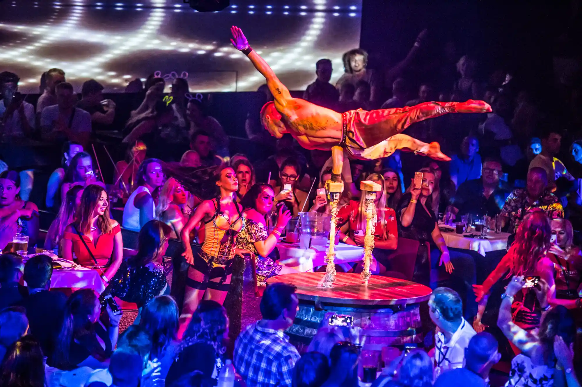 An acrobat balances on poles above the audience during a spectacular nightlife show with vibrant lighting.