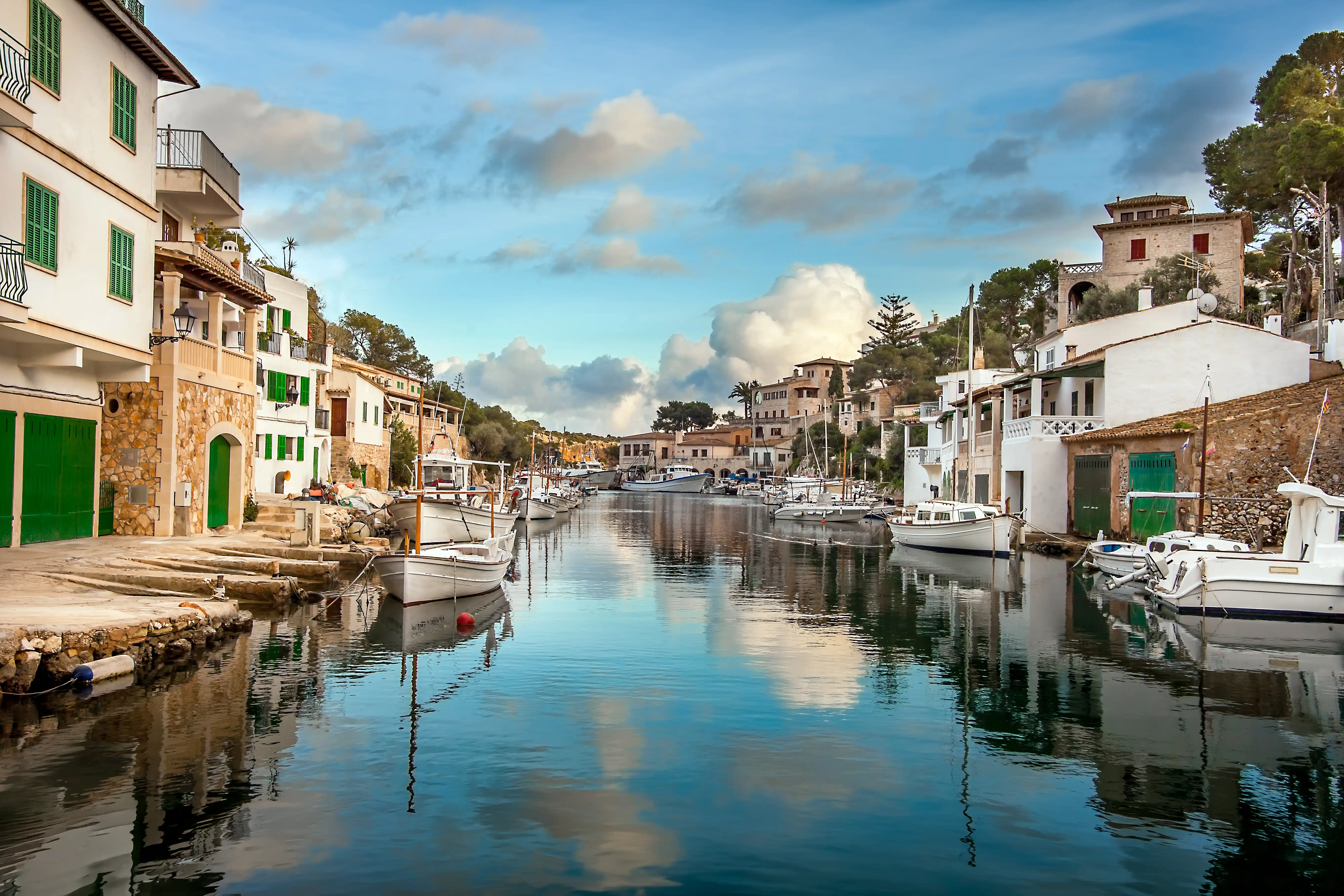 A picturesque fishing village harbor with traditional white houses, green shutters, and boats moored along the calm water channel.