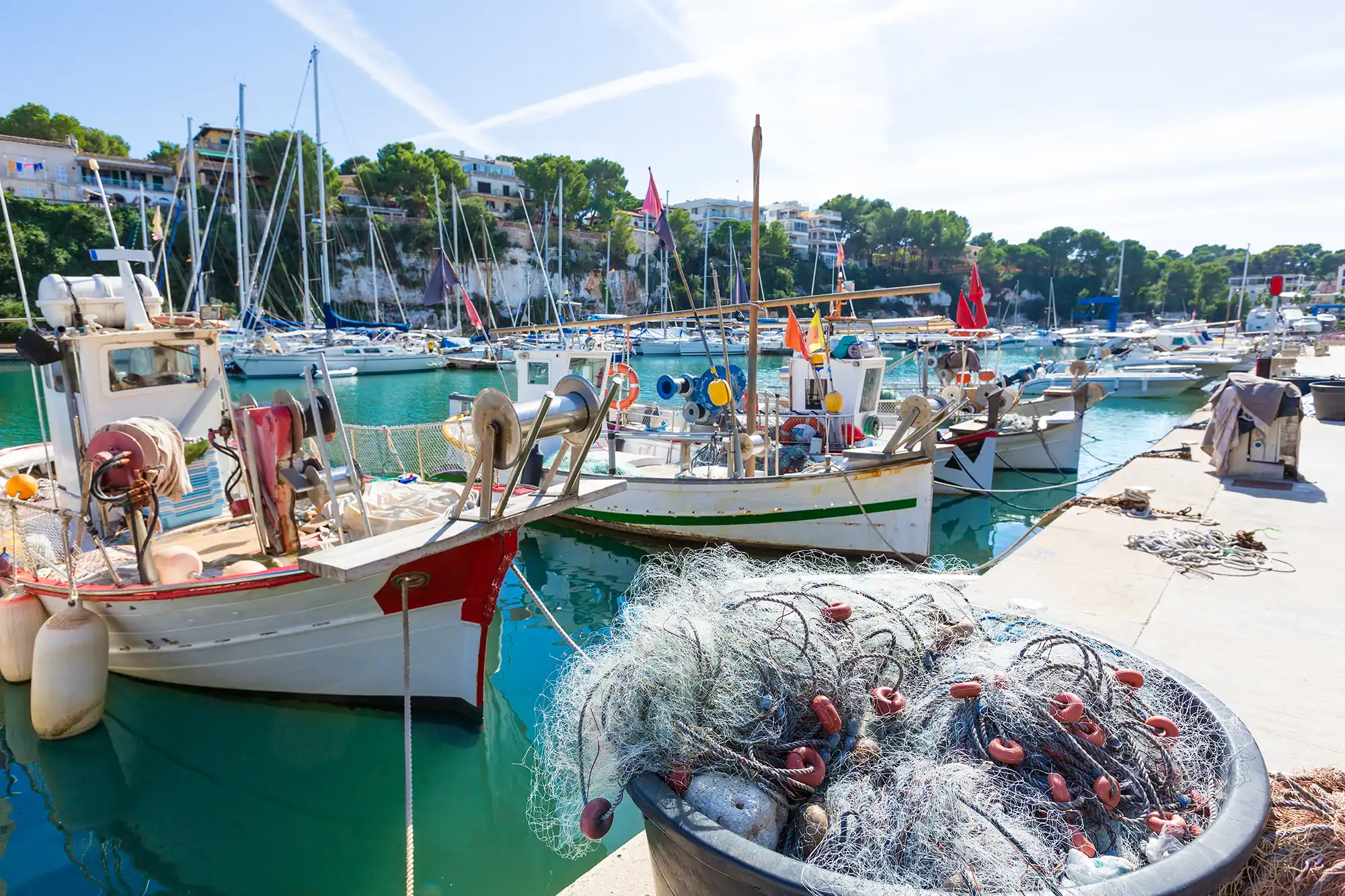 Traditional fishing boats are docked in a scenic harbor with a pile of fishing nets in the foreground.
