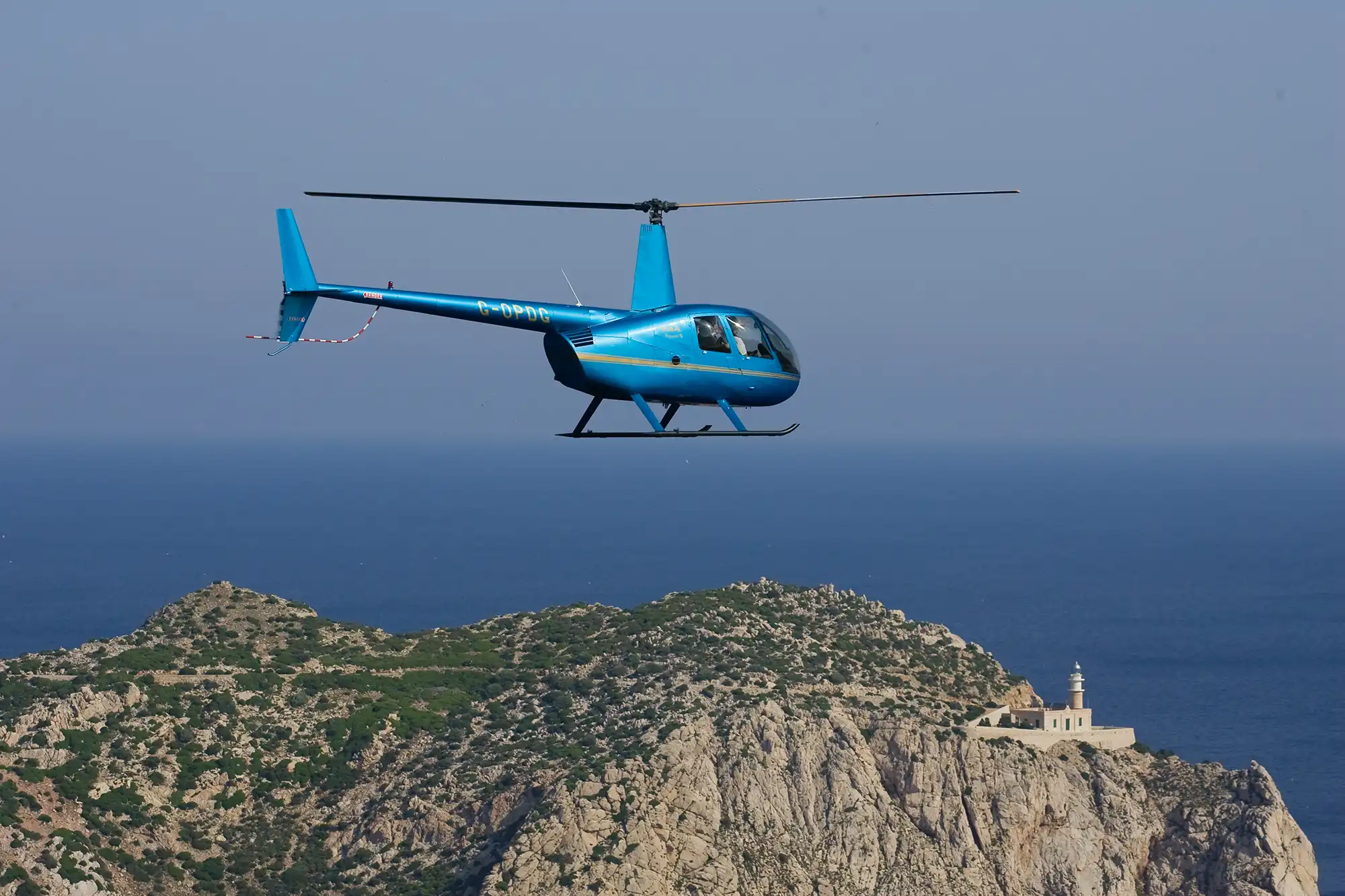 A blue helicopter flying in the air above a rocky coastal hill with a lighthouse and the sea in the background.