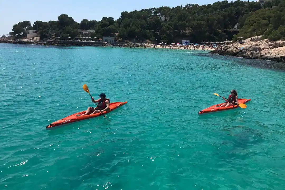 Two people paddle orange kayaks in vibrant turquoise sea water near a sandy beach surrounded by green trees.