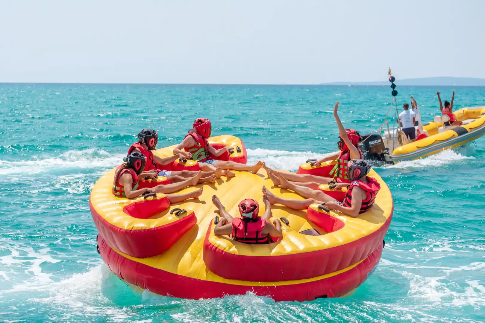 People wearing red helmets riding a circular inflatable towable boat on turquoise sea water.