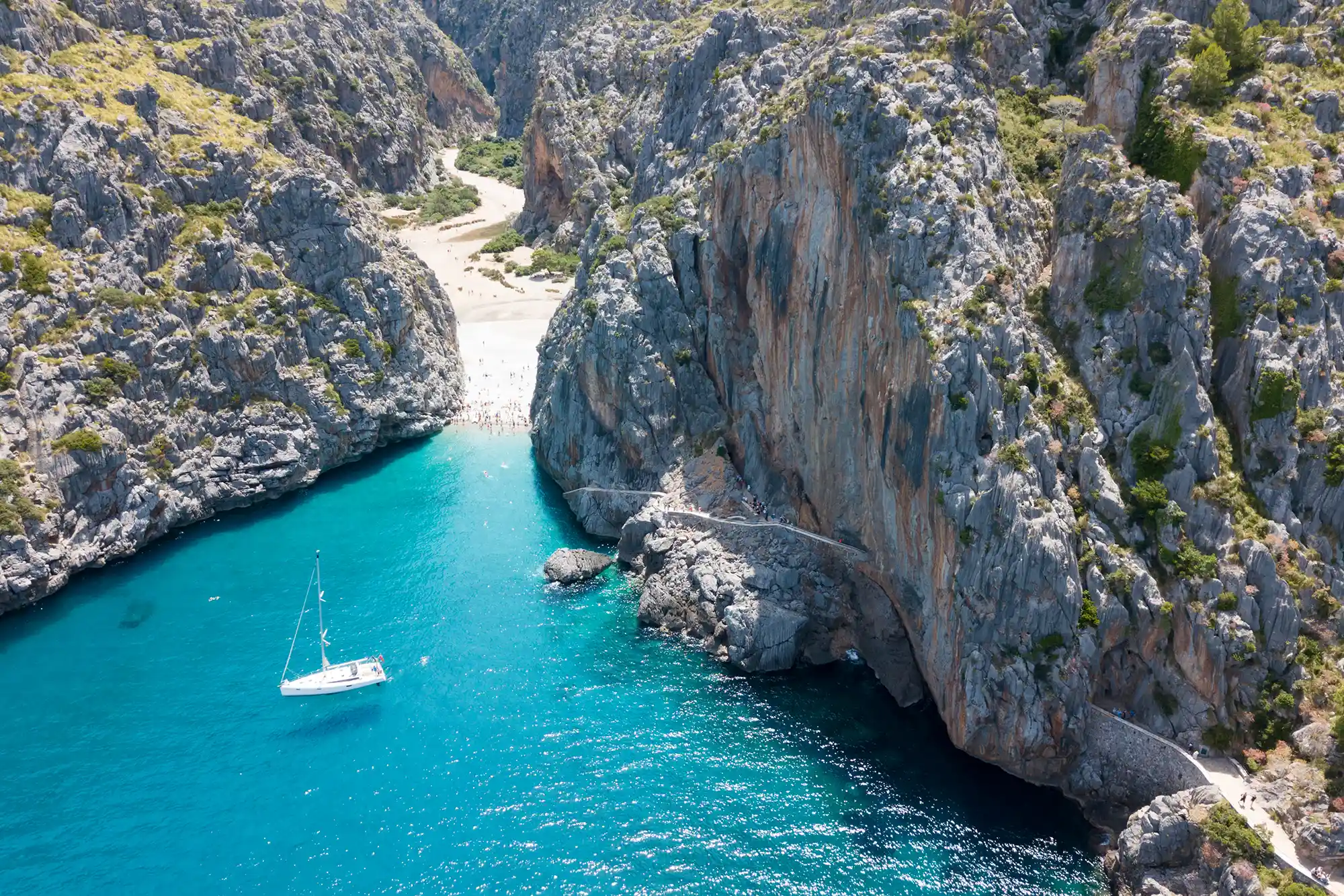 A high-angle view of a white sailboat in a turquoise bay between steep, rocky canyon walls leading to a beach.