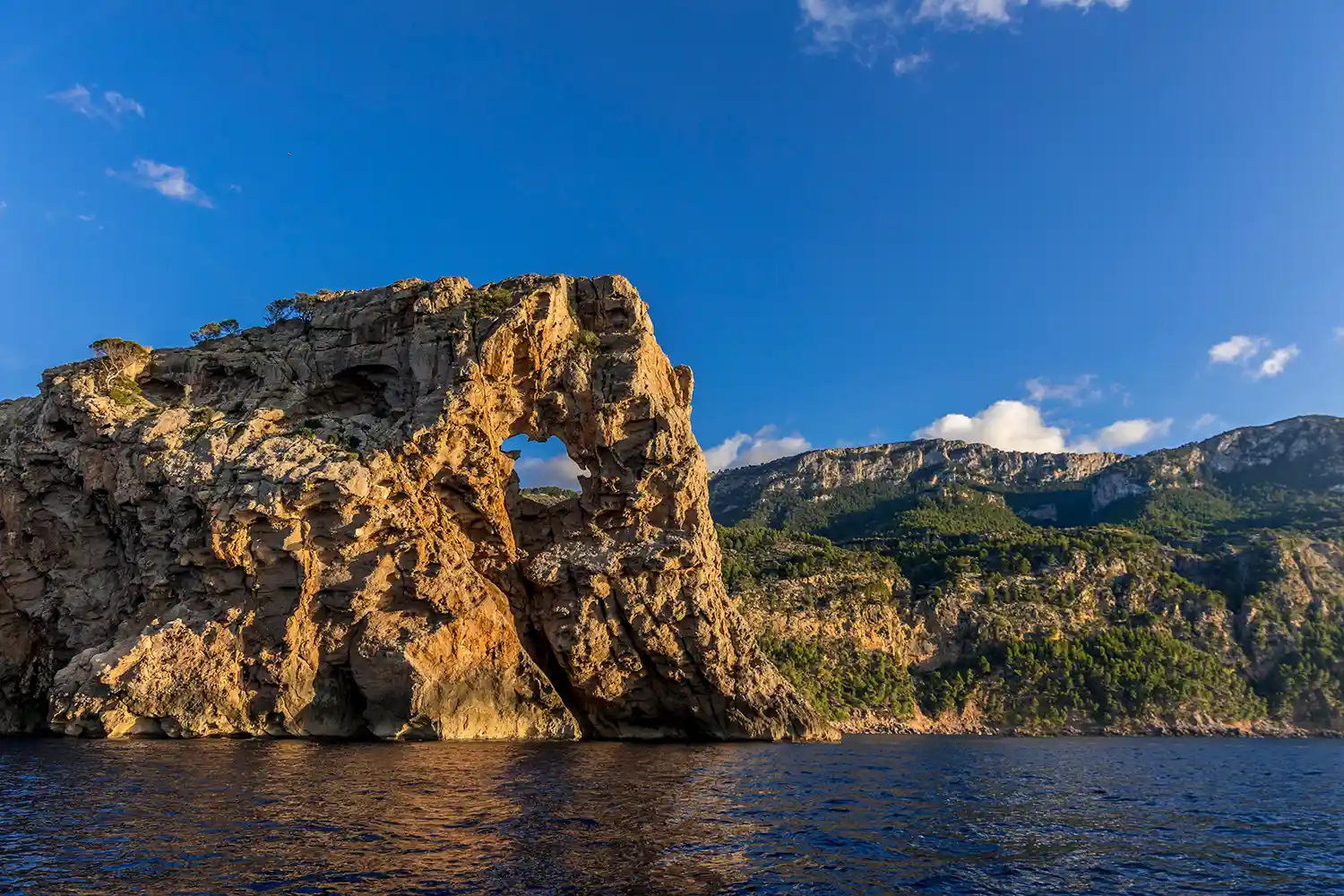 A majestic rock arch formation rising from the deep blue sea along a rugged coastline under a clear sky.