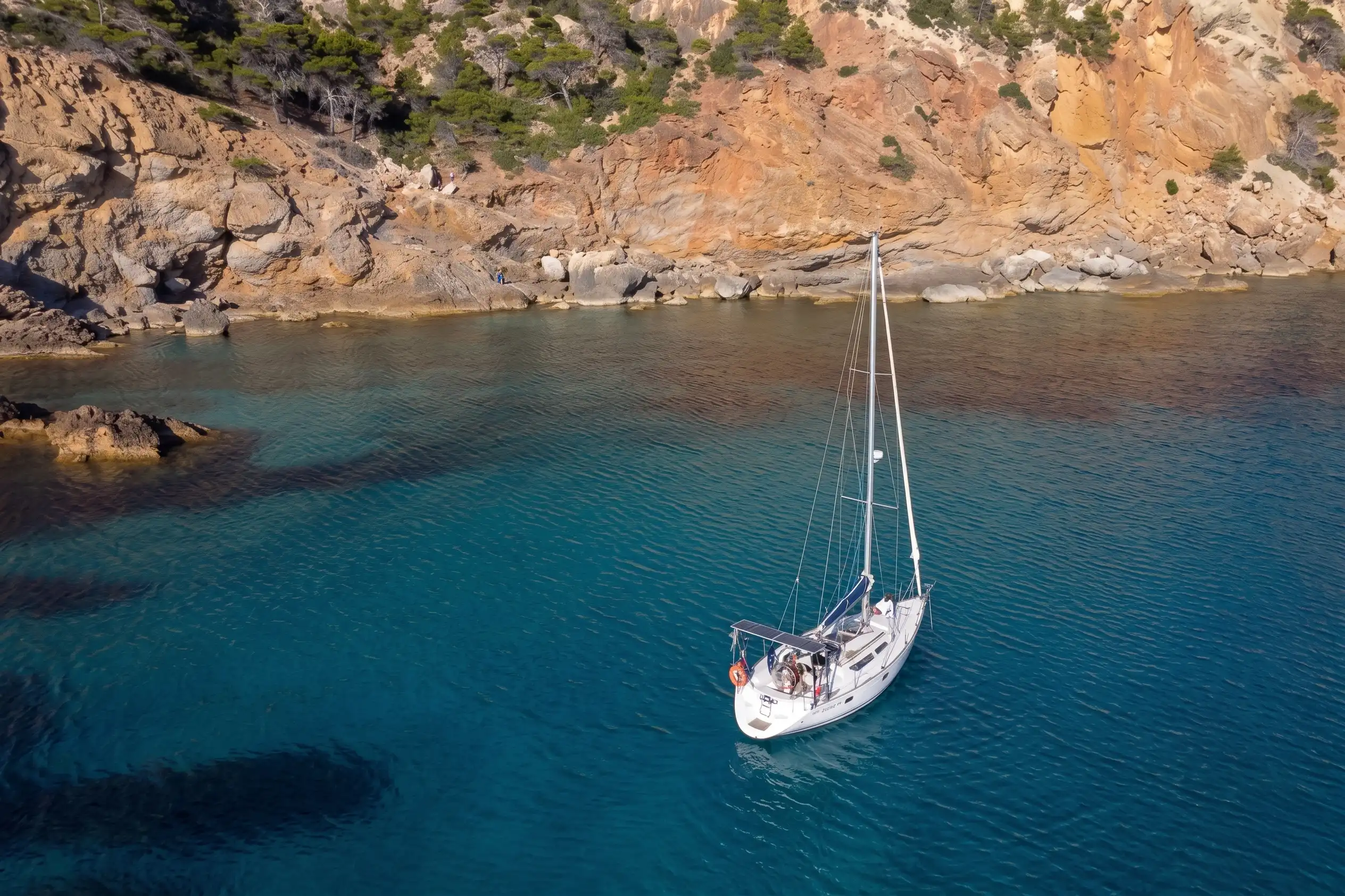 Aerial view of a white sailboat anchored in a quiet rocky bay with clear turquoise water.