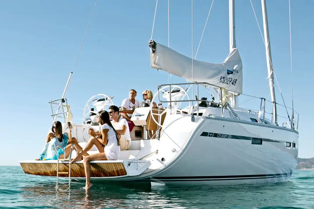A group of friends relaxes on the deck of a white sailboat on the calm blue sea under a clear sky.