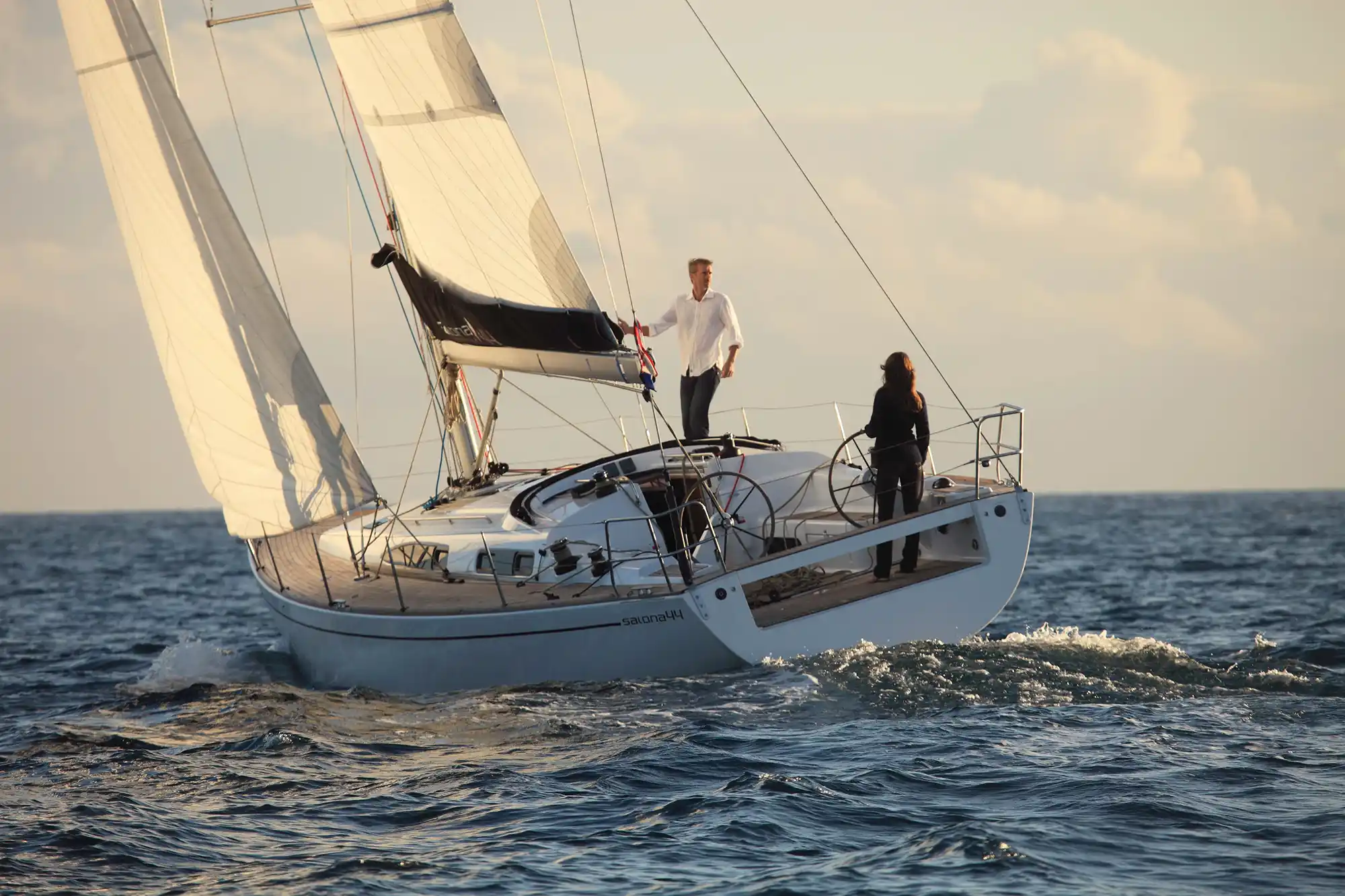 A man and a woman sail a white sailboat on the open sea under a warm, cloudy sunset sky.