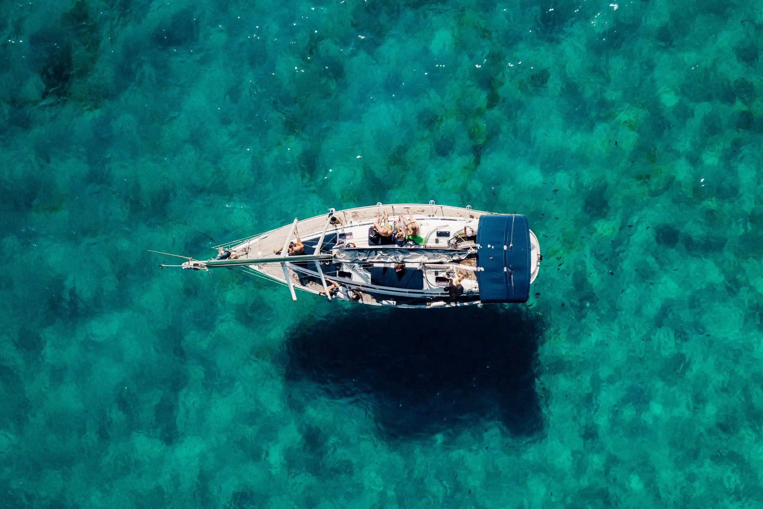 Vista aérea cenital de un velero flotando en aguas marinas turquesas de color cristalino, proyectando una sombra oscura sobre el fondo marino. Vista aérea cenital de un velero flotando en aguas marinas turquesas de color cristalino, proyectando una sombra oscura sobre el fondo marino.