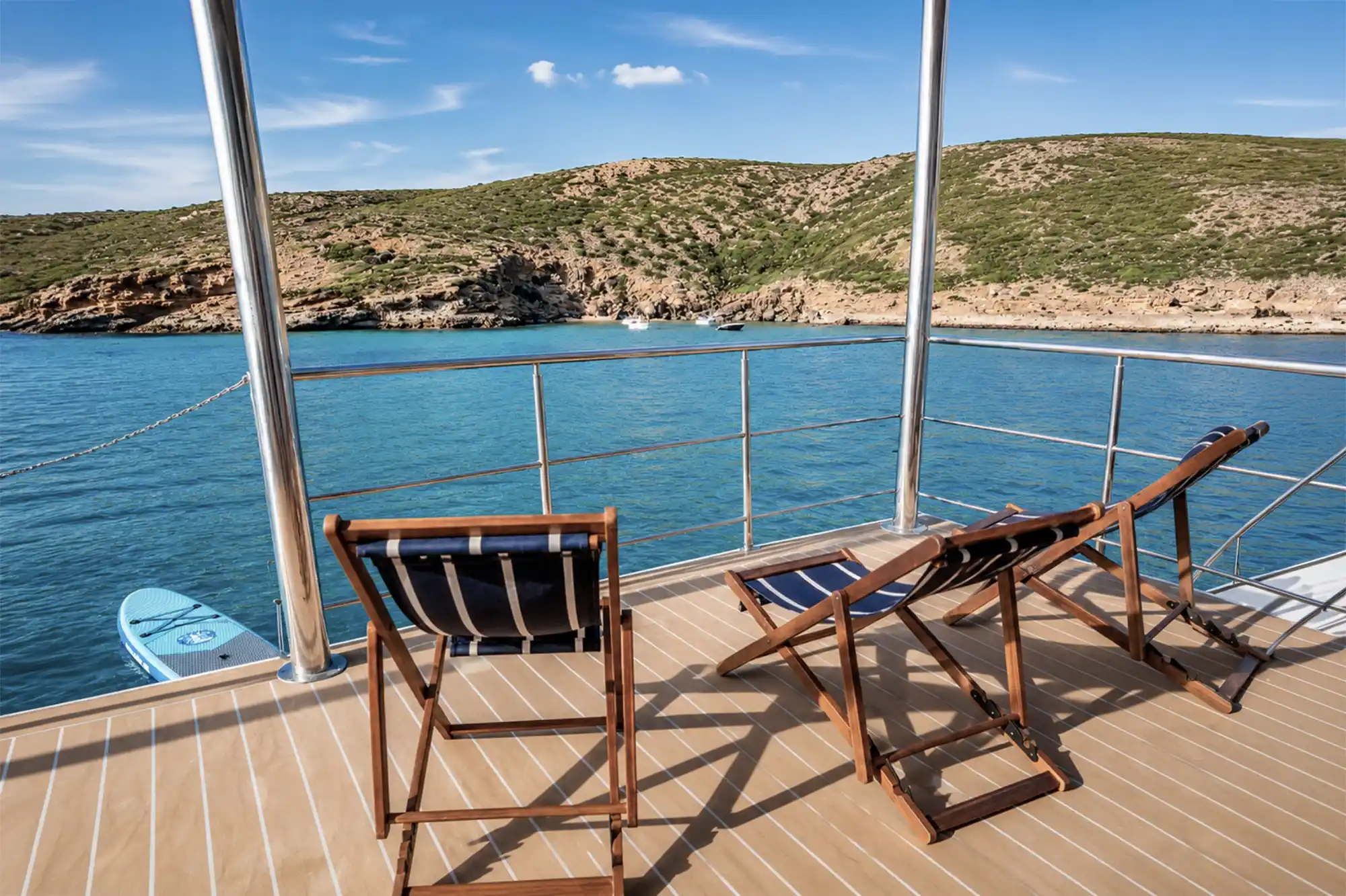Three wooden deck chairs sit on a boat platform overlooking a turquoise cove with hills under a bright blue sky.