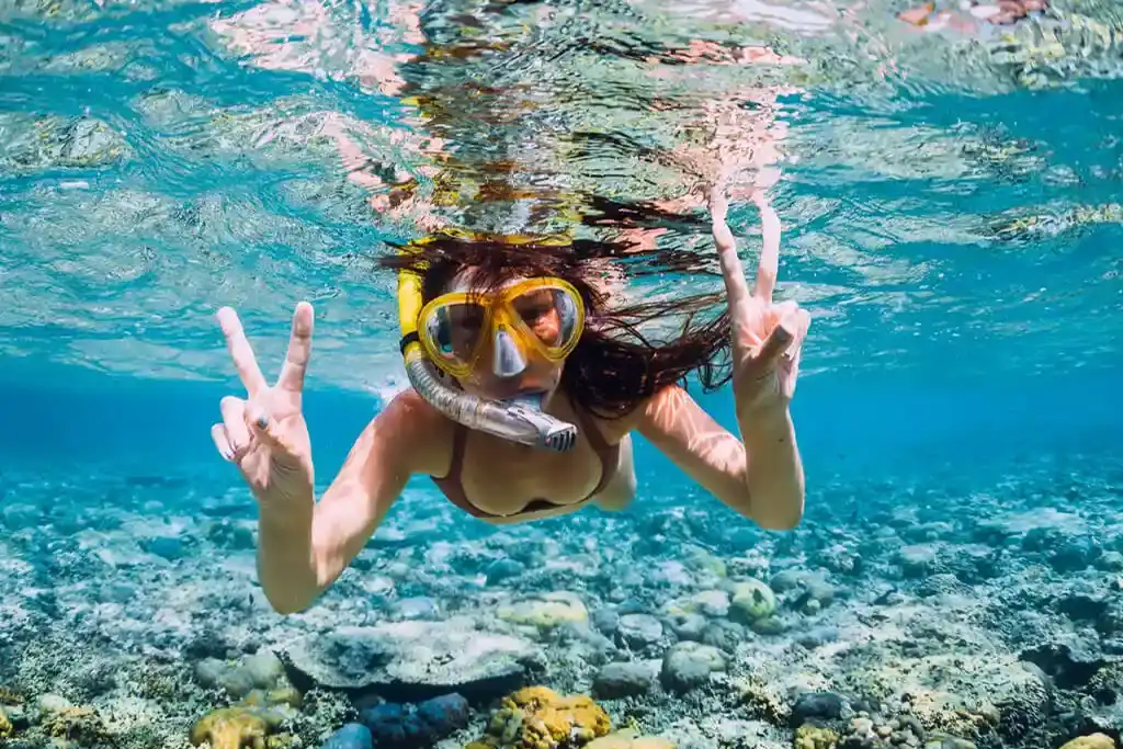 Eine Frau schnorchelt in klarem Wasser über einem Korallenriff und macht das Friedenszeichen.
