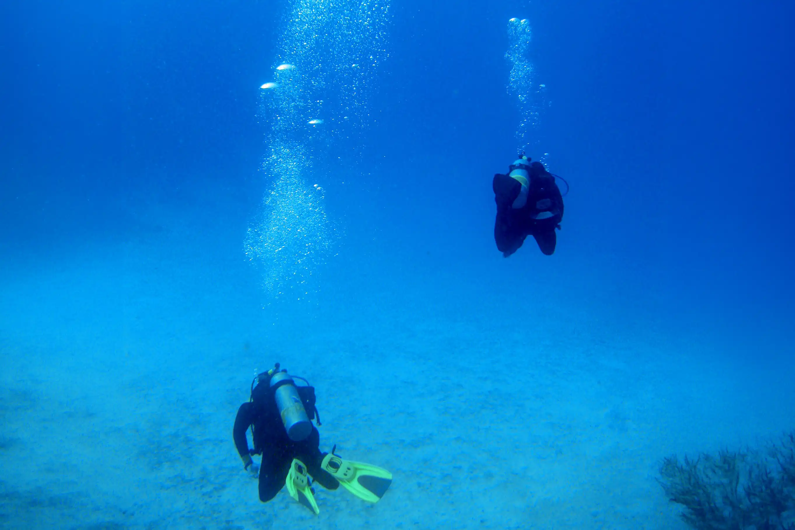Two scuba divers swimming deep in the blue ocean with air bubbles rising to the water surface.