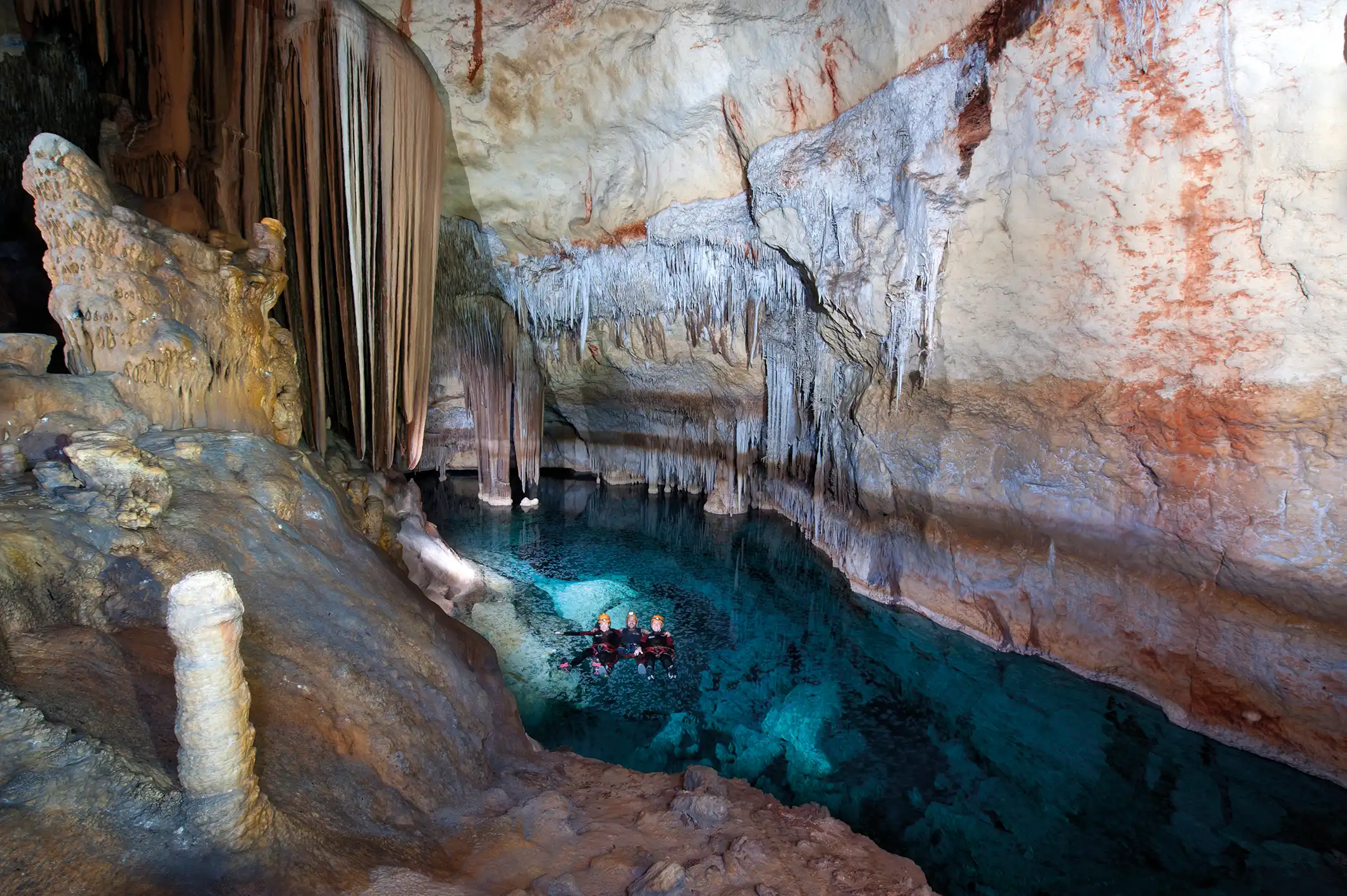 Scuba divers exploring an underground clear water lake surrounded by stalactites in a large stone cave.