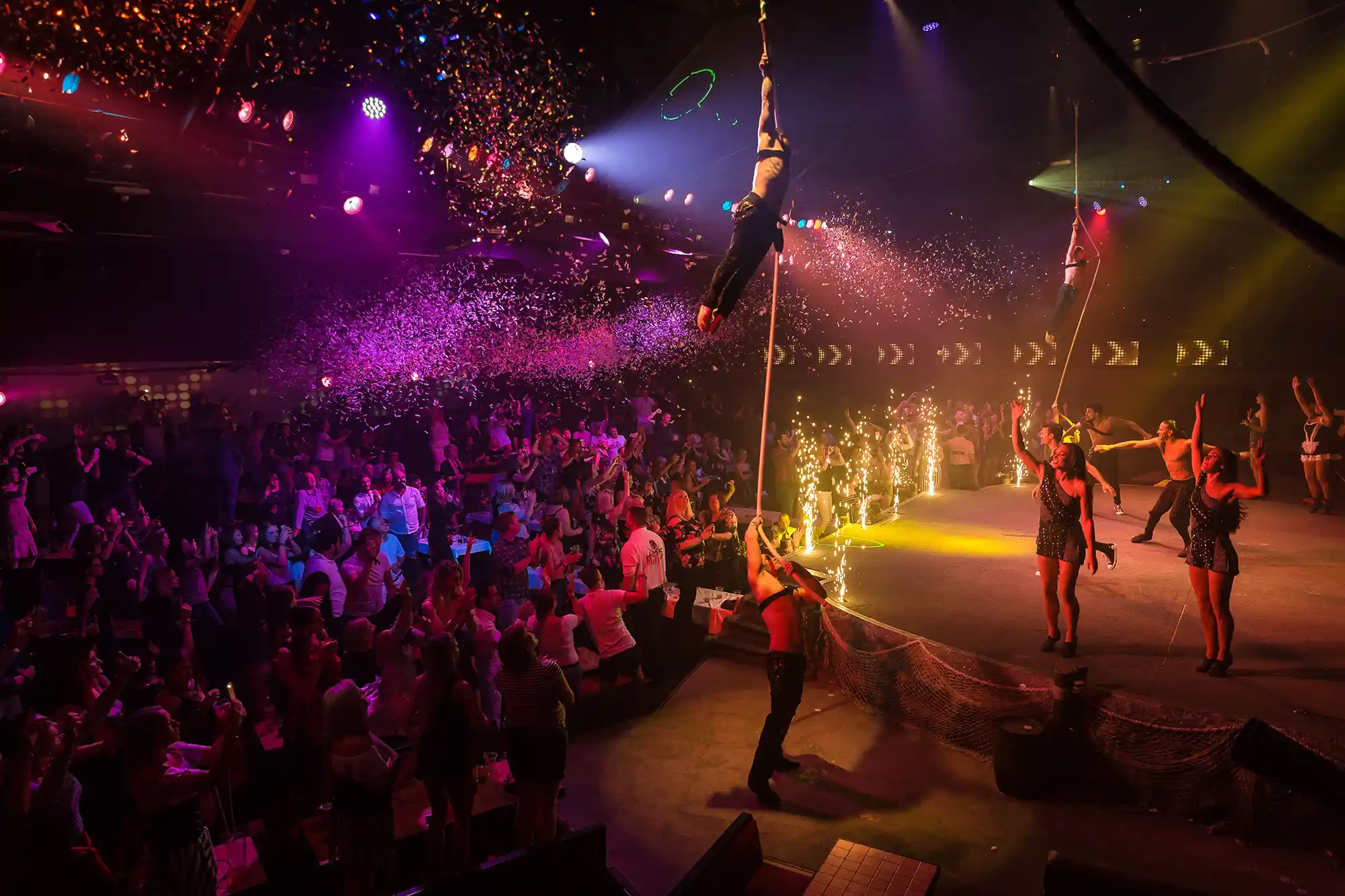 Aerial acrobats perform on ropes above a cheering crowd and dancers during a spectacular nightlife party show.