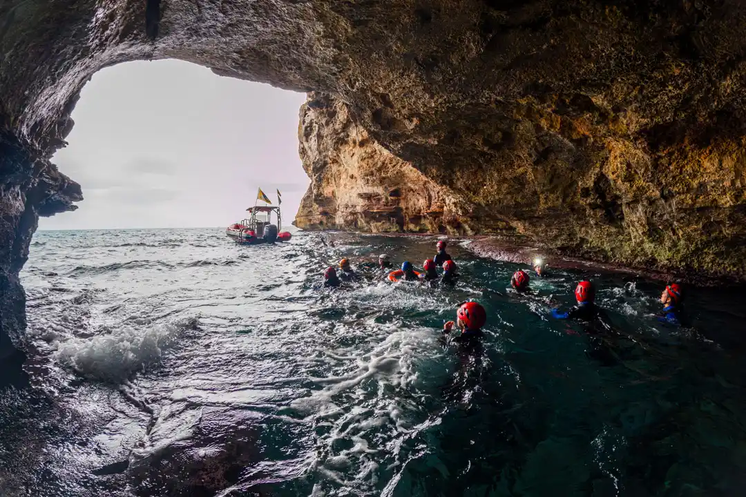 A group of swimmers wearing red helmets explores a sea cave, with a small boat visible at the cavern opening.