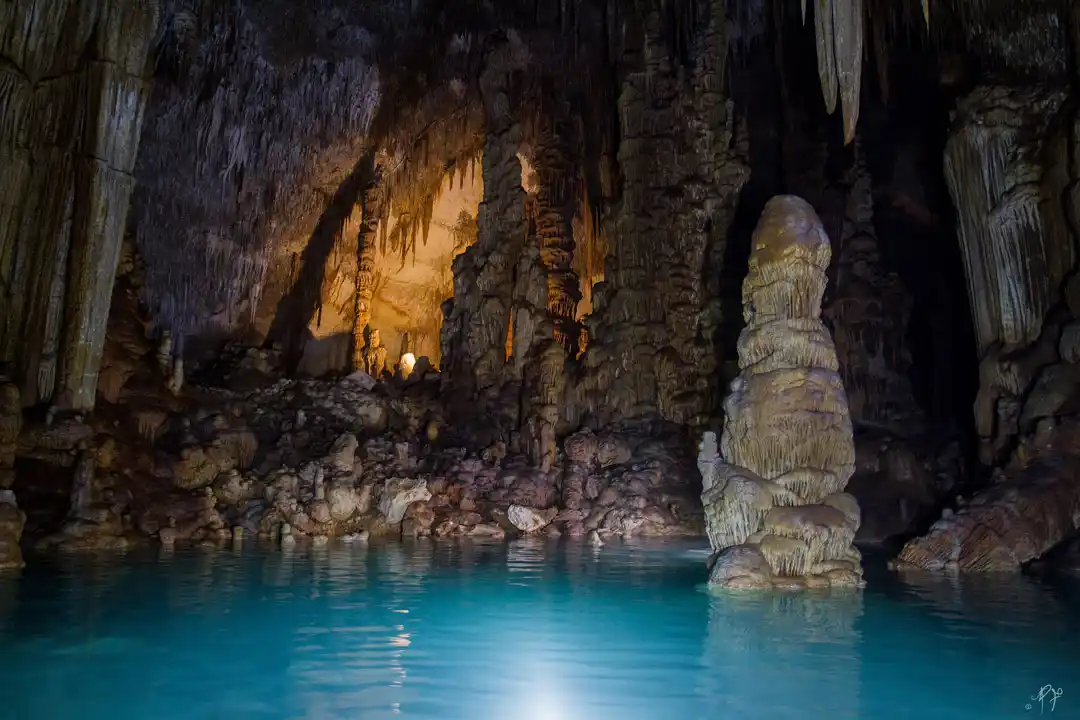 An underground cave interior featuring dramatic stalactites, stalagmites, and a clear turquoise water pool.