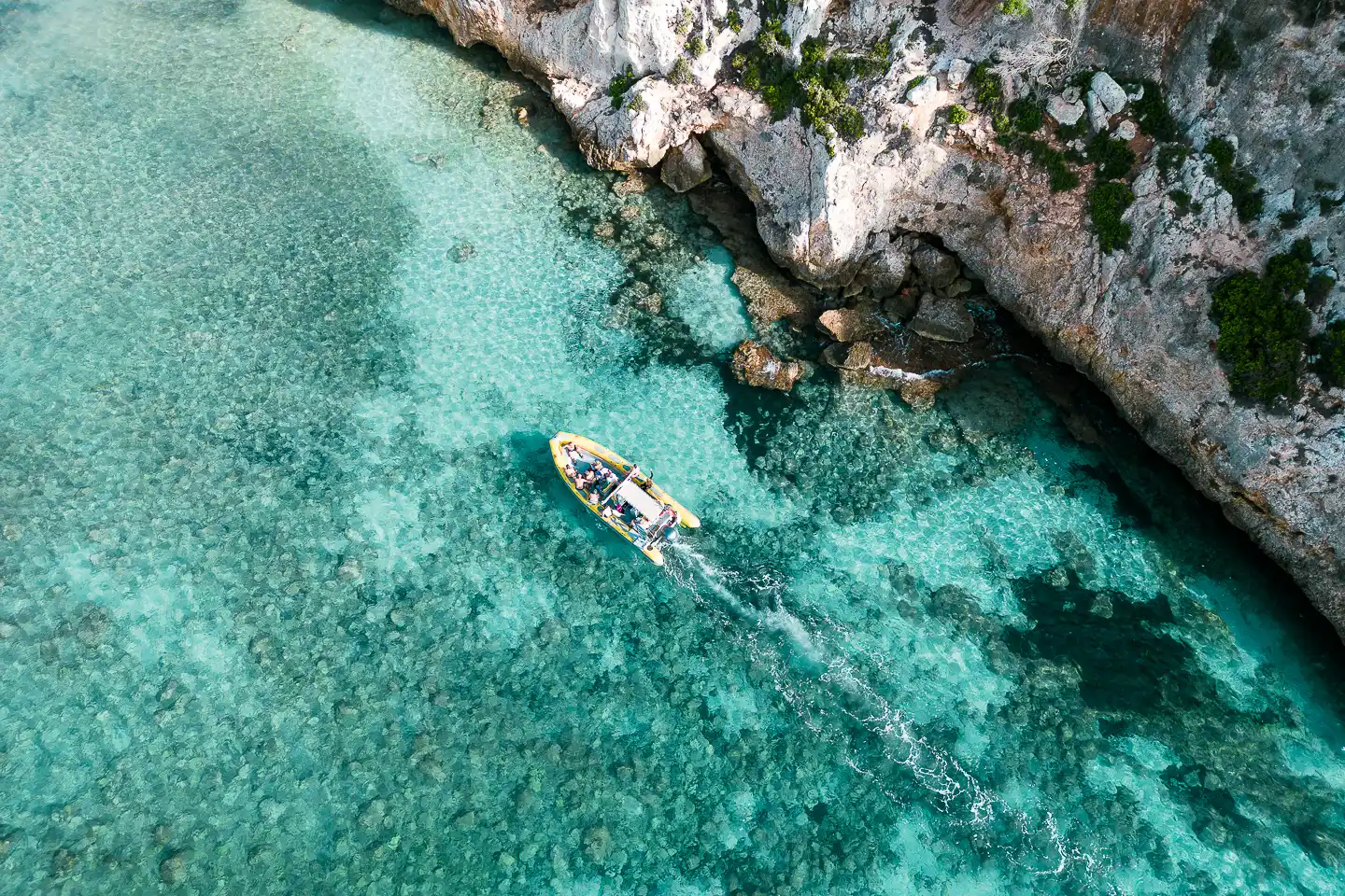 An aerial top-down view of a small motorboat cruising through crystal-clear turquoise waters next to rocky cliffs.