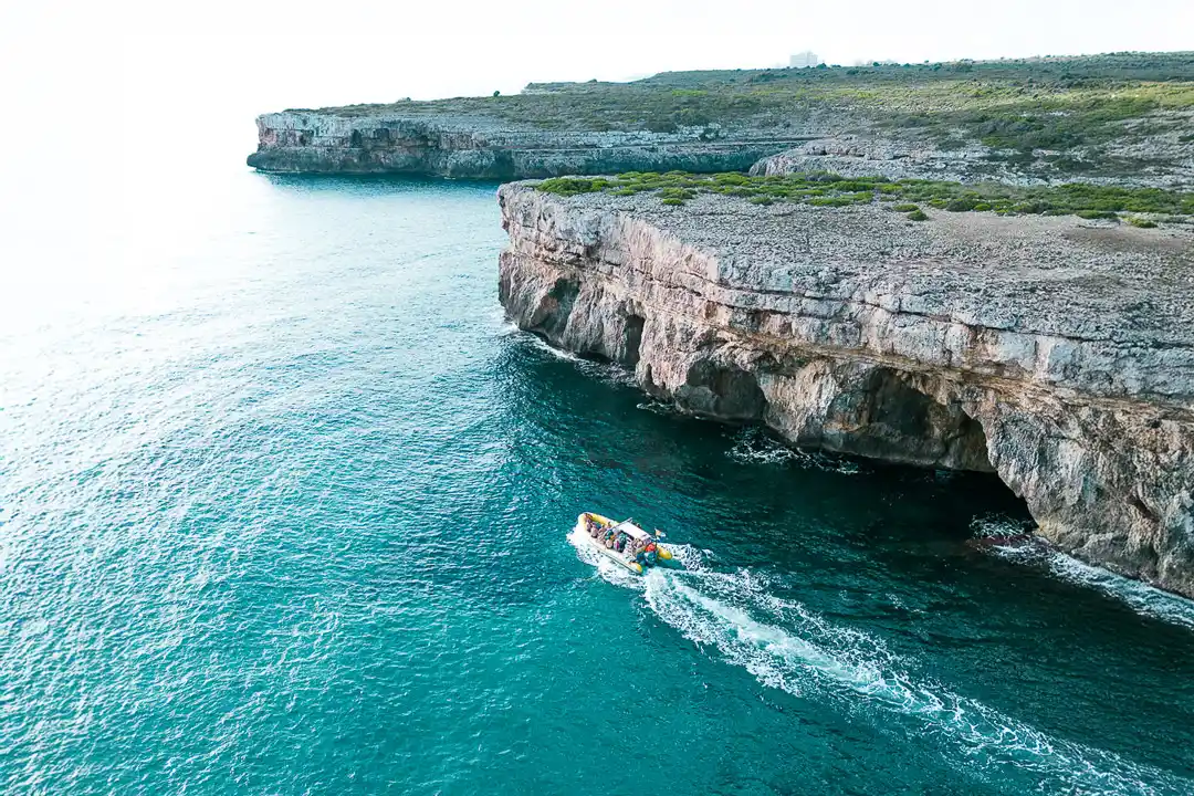 An aerial view of a motorboat cruising next to dramatic rocky cliffs along a bright turquoise coastline.