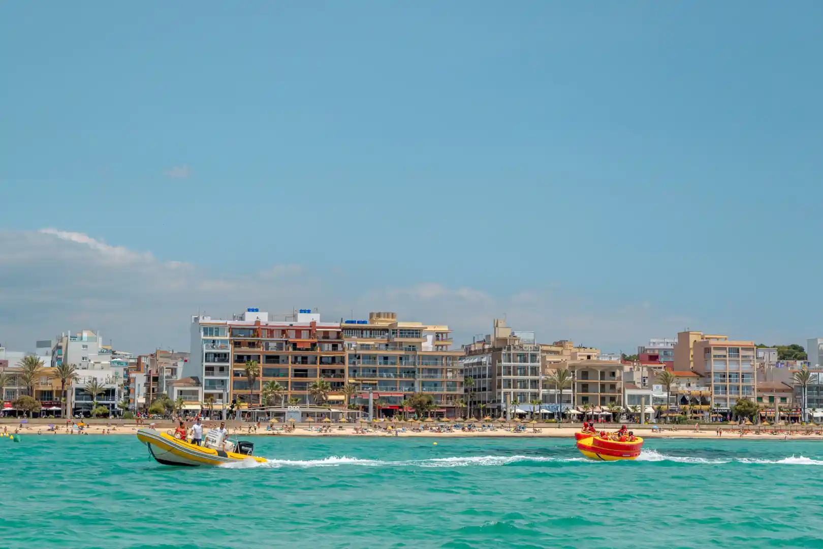 Zwei kleine Schnellboote fahren auf türkisfarbenem Meer vor einem Küstenstrand mit Hotels im Hintergrund.