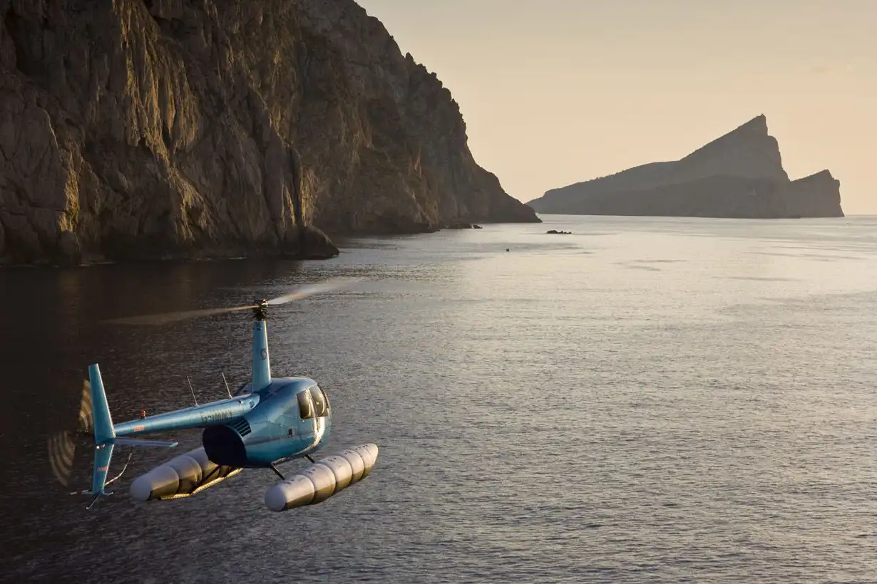 A blue helicopter equipped with floats flying over the sea near a steep rocky cliff during a golden sunset. A blue helicopter equipped with floats flying over the sea near a steep rocky cliff during a golden sunset.