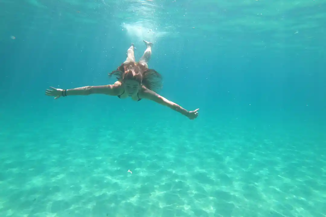 A young woman swims underwater with her arms outstretched in a clear turquoise sea with a sandy seabed.