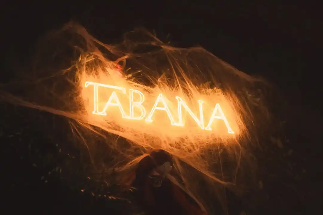 Neon sign reading TABANA with a warm orange glow is draped in artificial spiderwebs against a dark, atmospheric background.